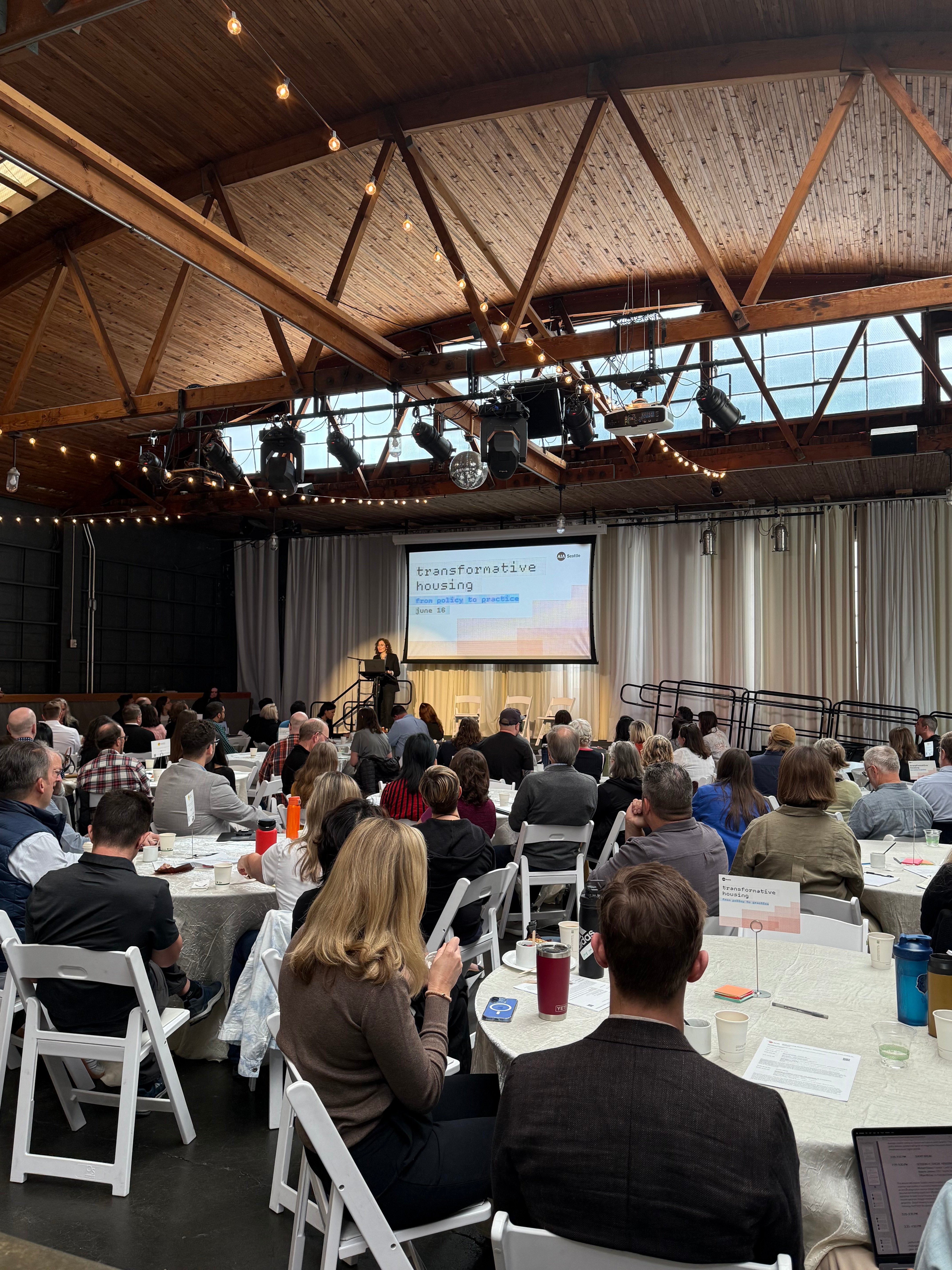 Audience seated at round tables listening to a speaker on stage under a wood-beamed ceiling with string lights, at a 'Transformative Housing' forum