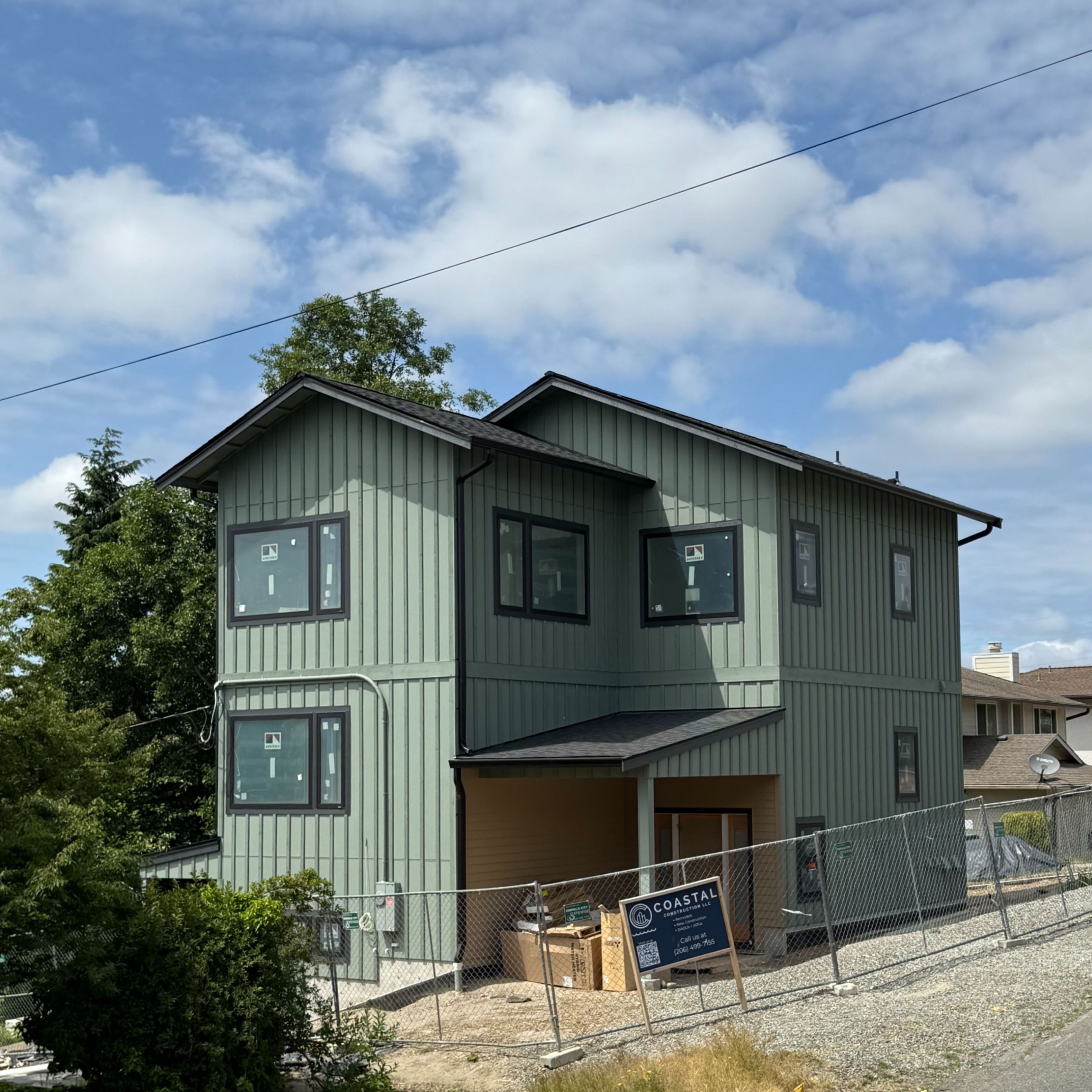 Exterior of a three-story house under construction, with green vertical siding installed, black-trimmed windows, and a construction fence in front