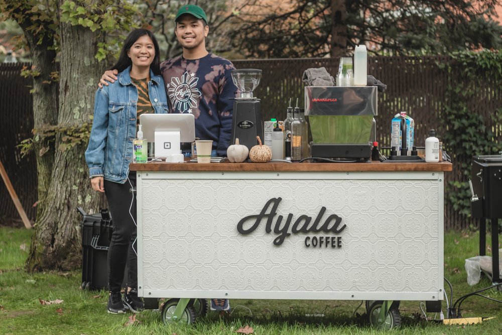 Two people smiling behind a mobile Ayala Coffee cart set up outdoors on grass, surrounded by trees