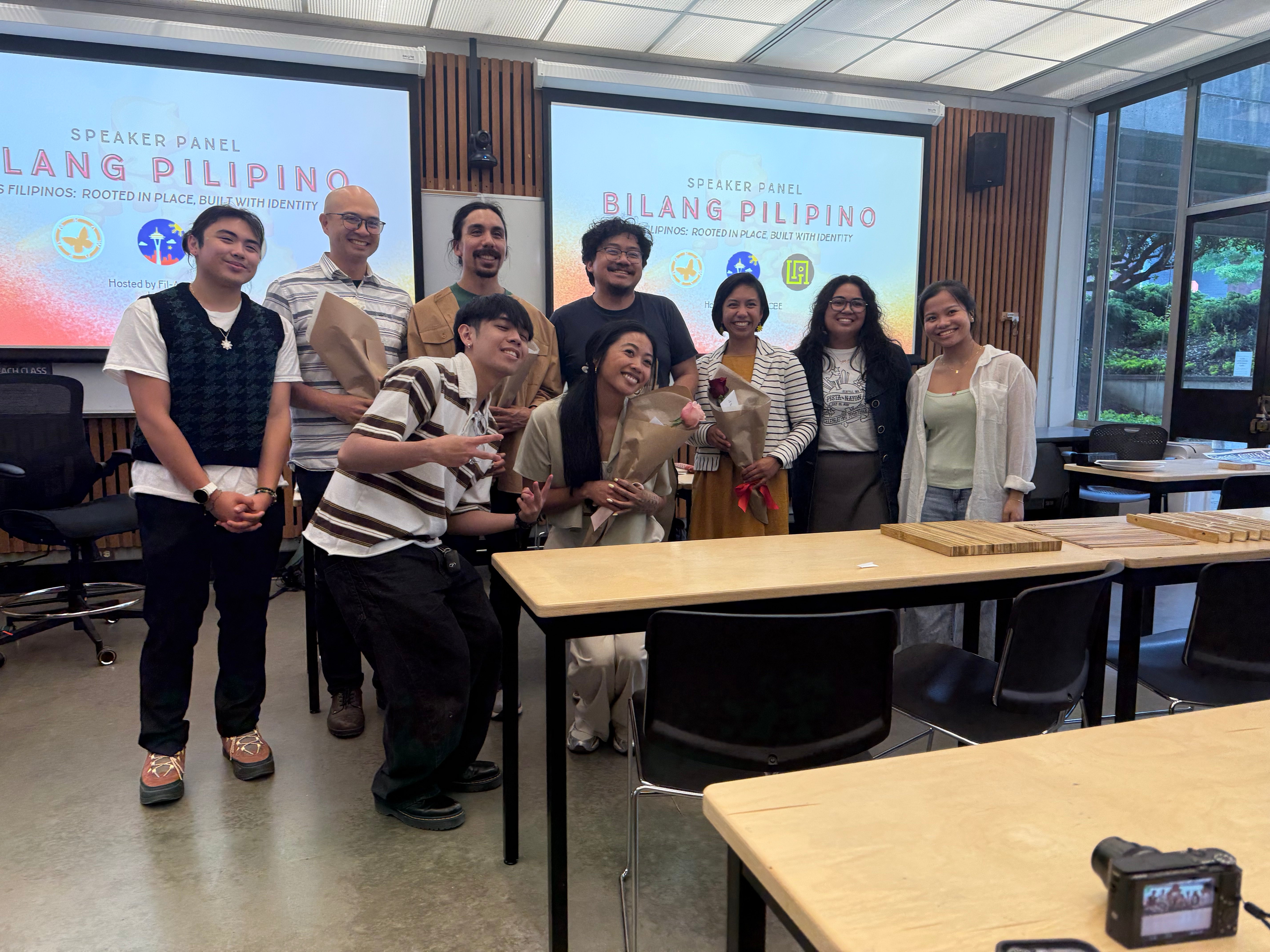 Group of nine people posing and smiling together in a classroom, with two 'Bilang Pilipino' speaker panel screens displayed behind them