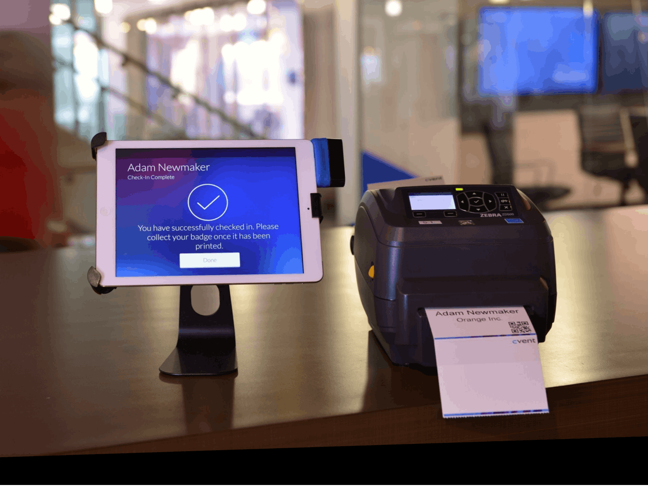 An event check-in kiosk showing a 'Check-In Complete' confirmation on a tablet, with a badge printer beside it printing an attendee's name badge