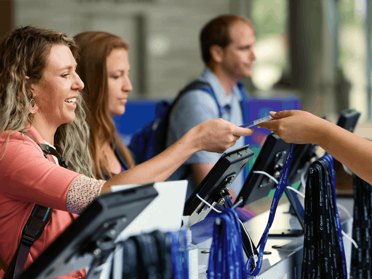 Event check-in staff handing a badge to an attendee at a registration desk, with lanyards and tablets visible in the background