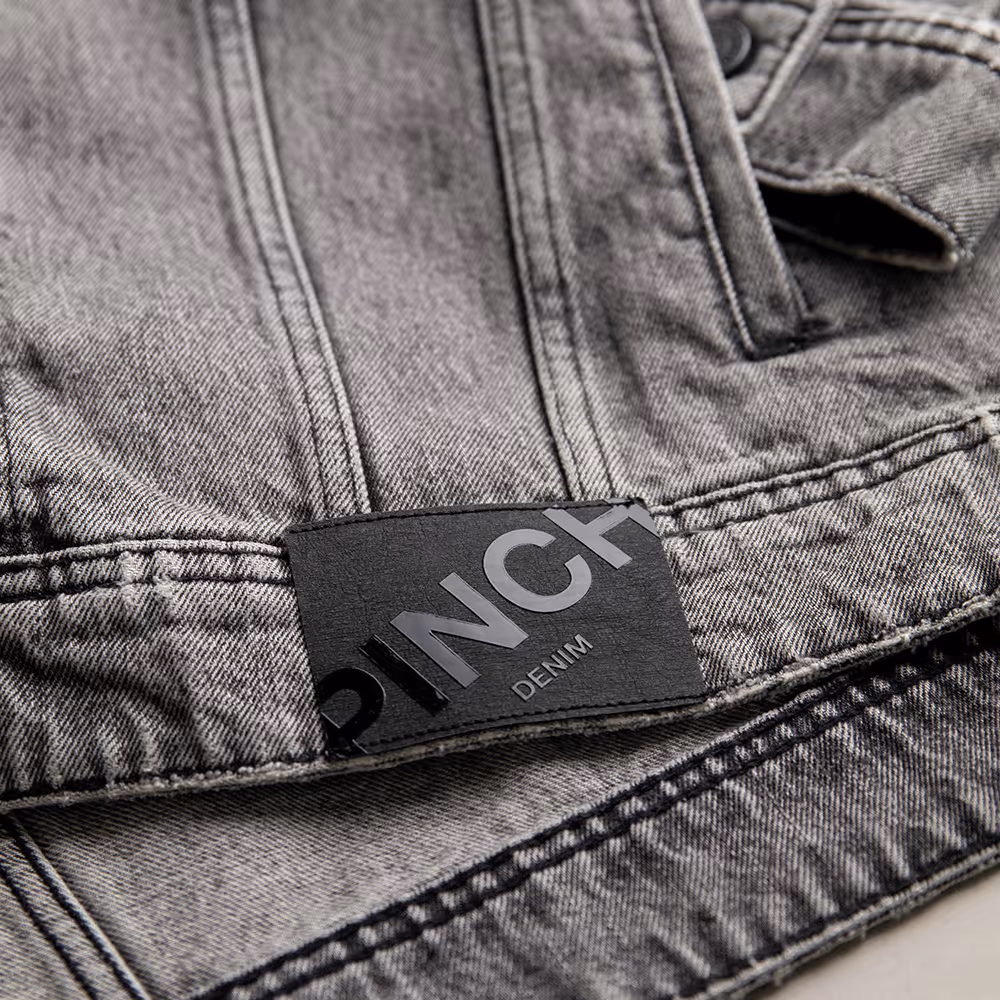 Close-up of a premium denim jacket featuring a glossy "Pinch" logo on a branded hem patch.