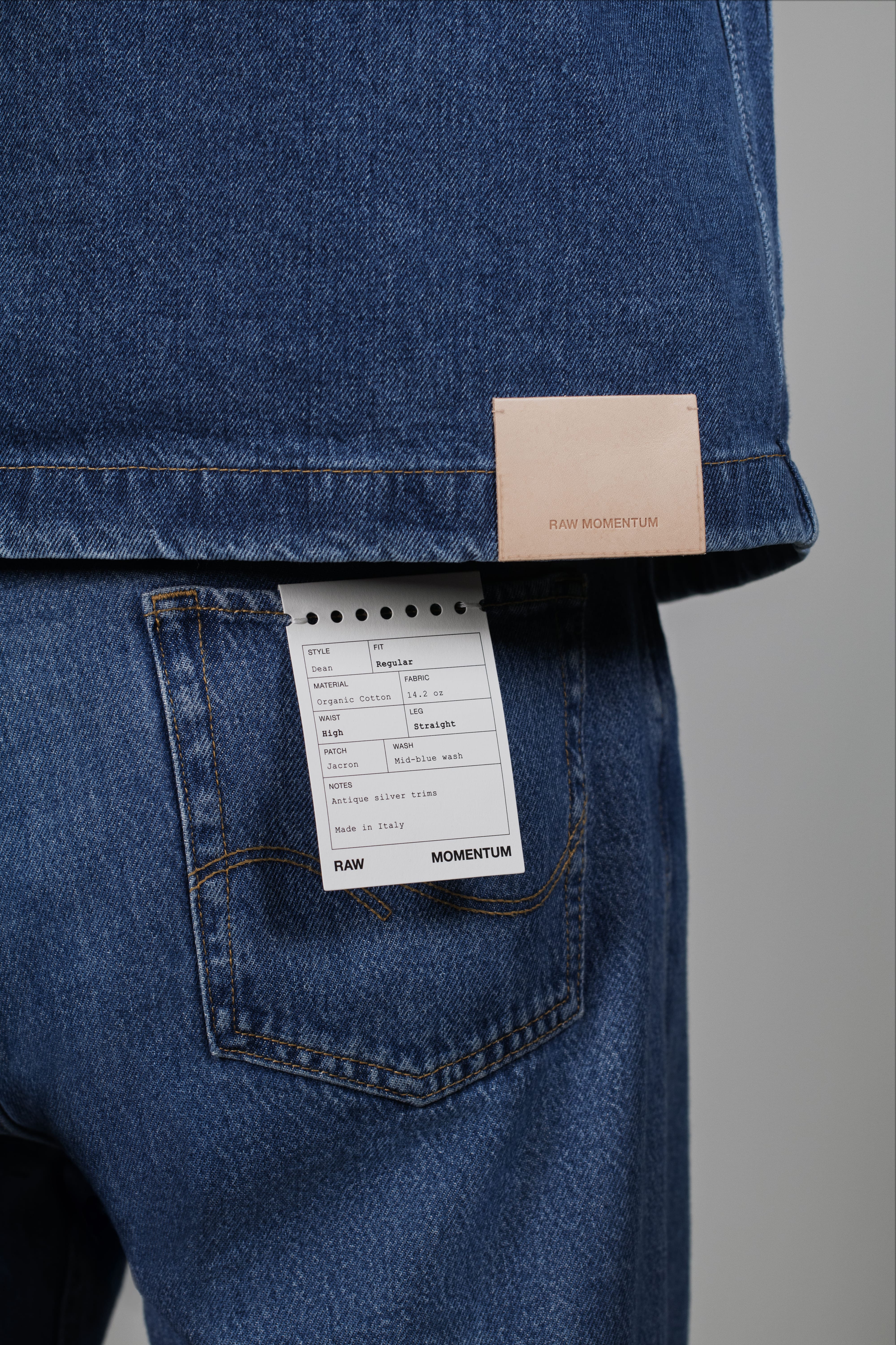 Model wearing a denim jacket with a branded hem patch and jeans featuring a branded back pocket label against a neutral background.