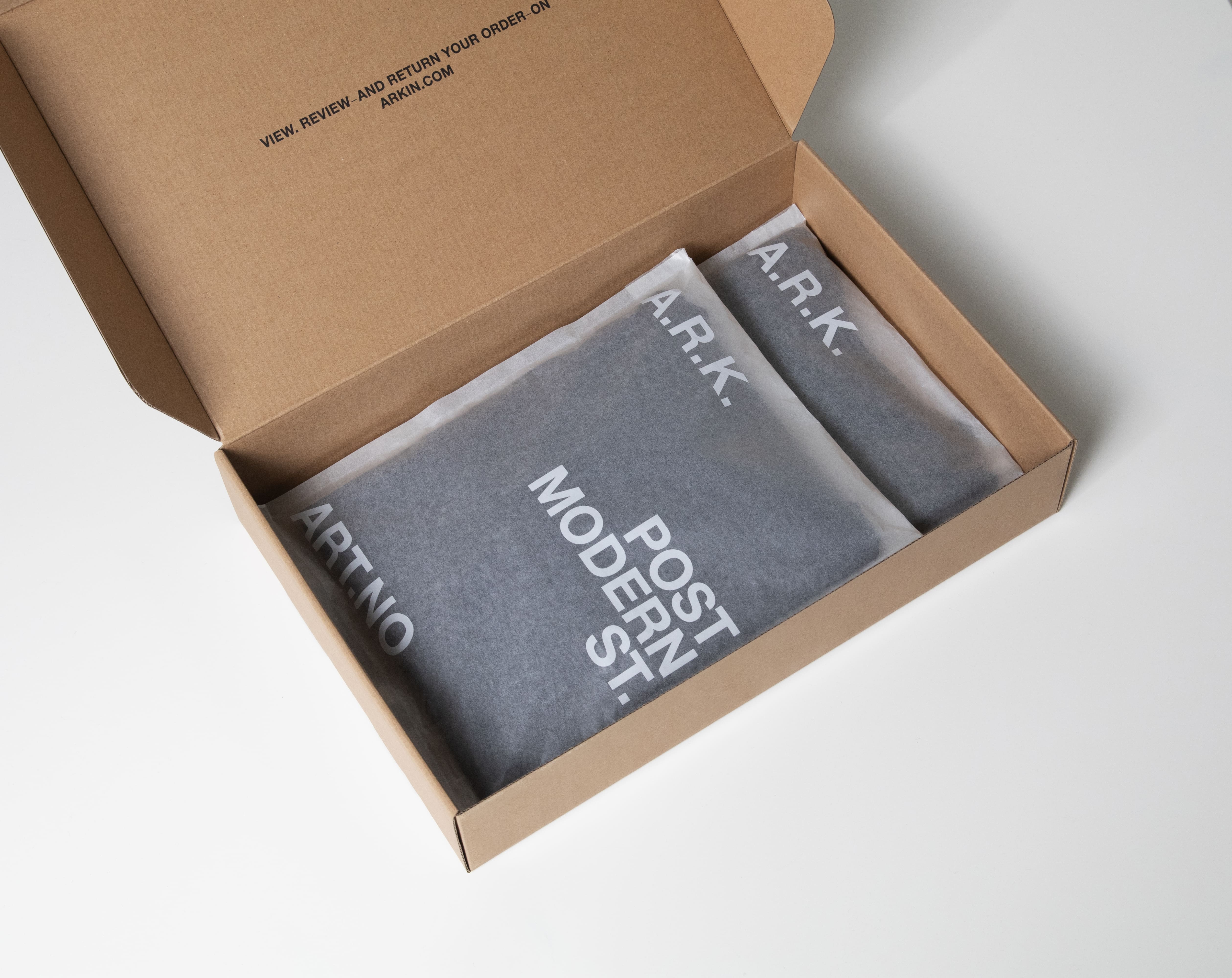 Open cardboard box containing two neatly folded garments wrapped in tissue paper, labeled with white tex