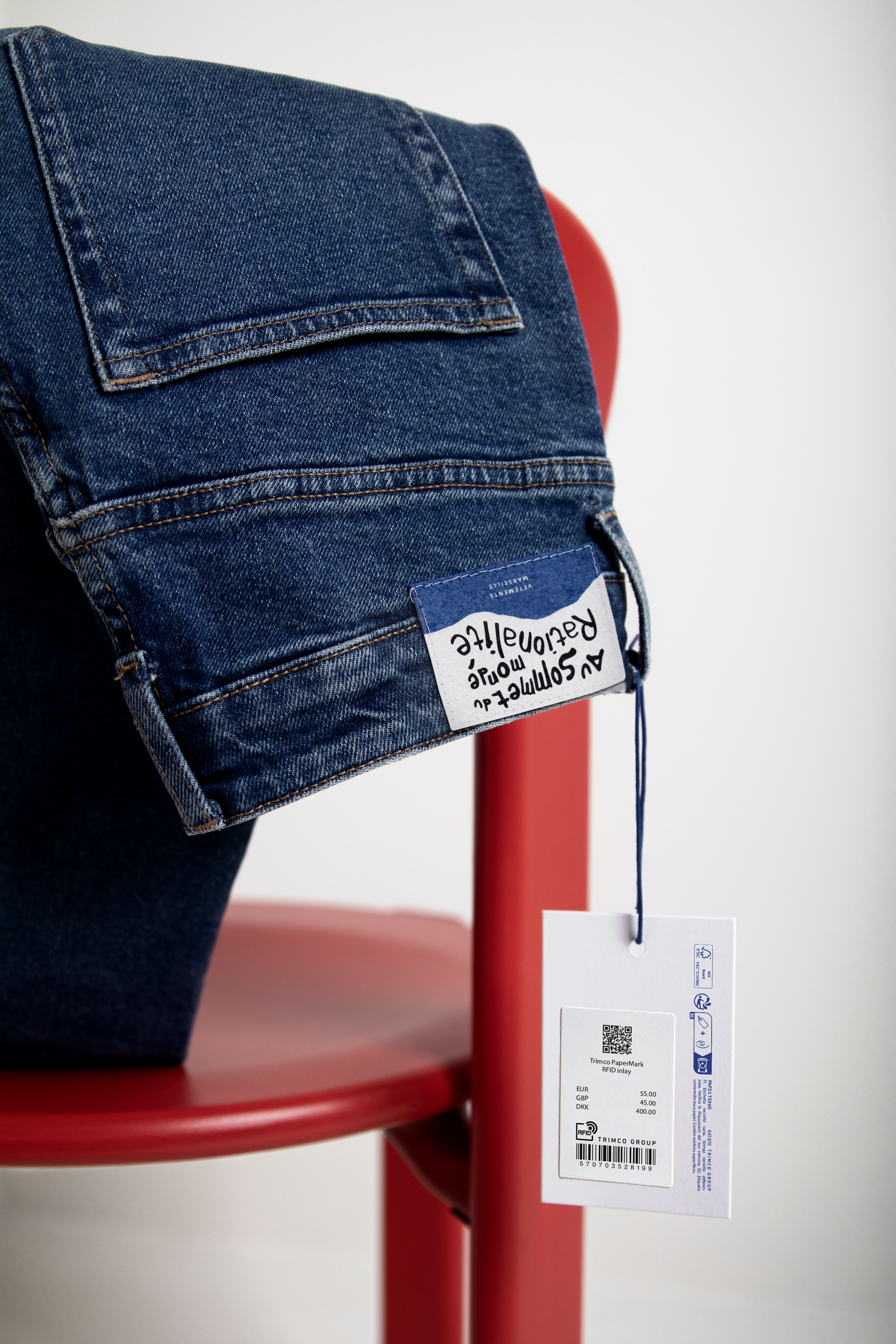 Folded blue jeans displayed on a red chair, featuring a branded back patch and RFID hangtag against a white background.