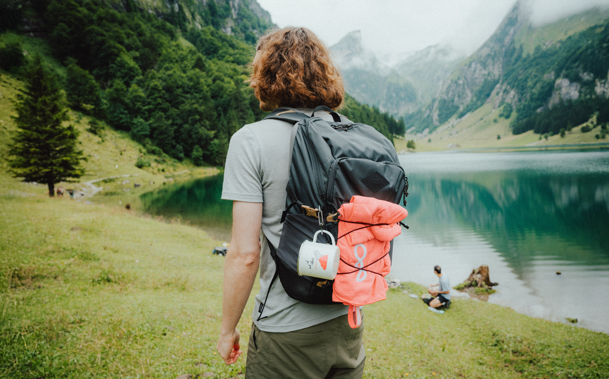 Eine Person, die neben einem Bergsee steht. Sie trägt einen Wanderrucksack, an dem eine Emaille-Tasse und ein Microfasertuch befestigt sind.