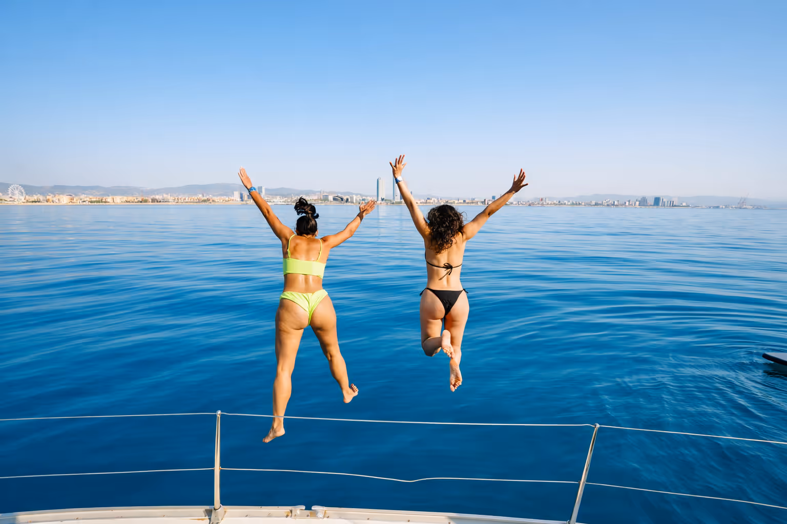 Two women smiling and relaxing on the deck of a sailboat near a harbor with buildings and yachts in the background.