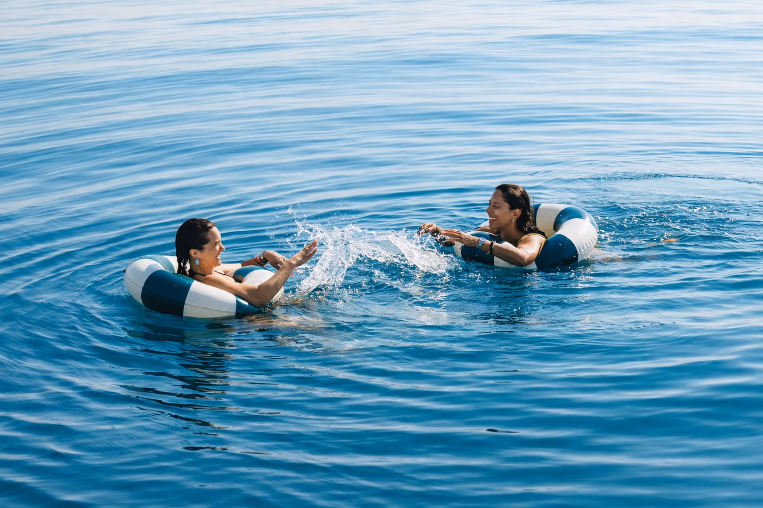 Two people floating in the calm ocean with life rings near the side of a boat under clear blue sky.