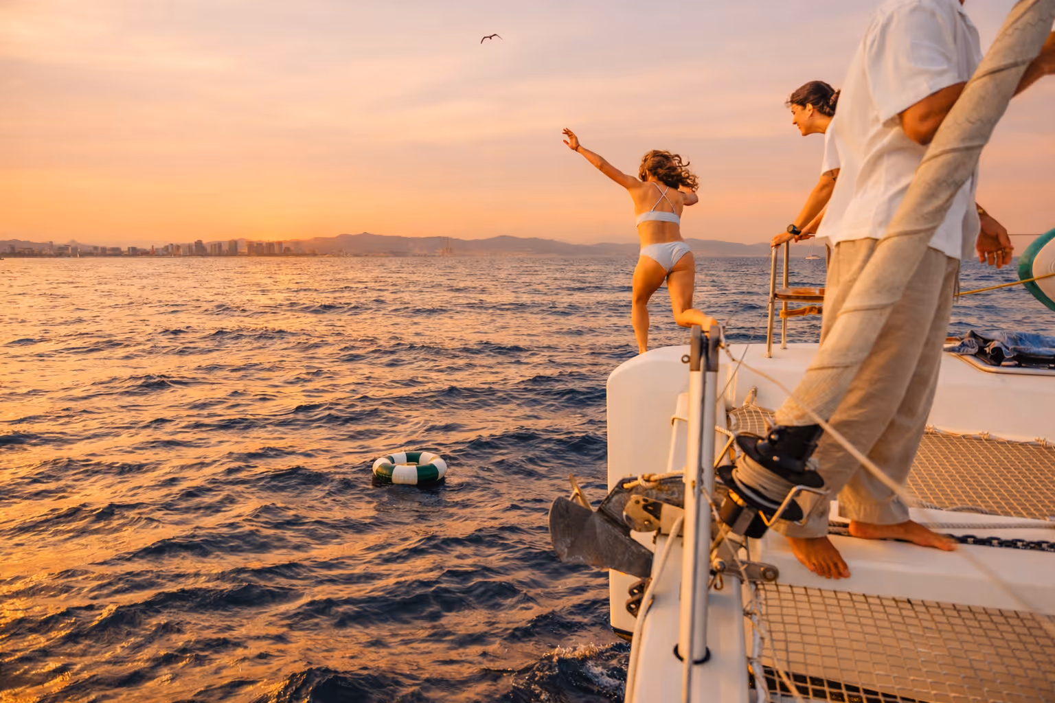 Woman in a white bikini jumping off the edge of a catamaran into the ocean at sunset.