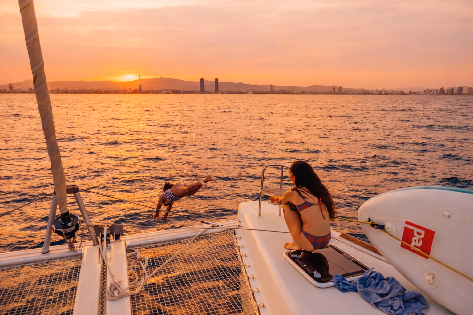Person in a bikini jumping off a catamaran into the ocean at sunset with others watching.