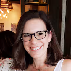 Smiling woman with shoulder-length brown hair and glasses in a warmly lit indoor setting.