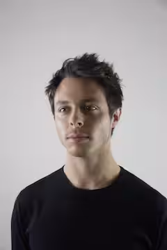 Portrait of a young man with dark hair wearing a black shirt against a plain light background.