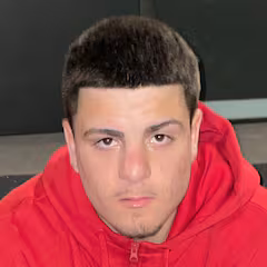 Close-up of a young man with short dark hair wearing a red hoodie looking directly at the camera.