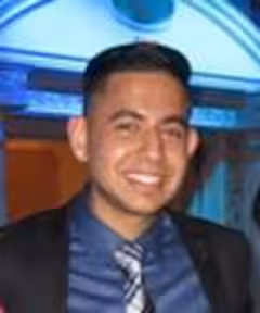 Smiling man with short dark hair wearing a dark suit, blue shirt, and striped tie indoors.