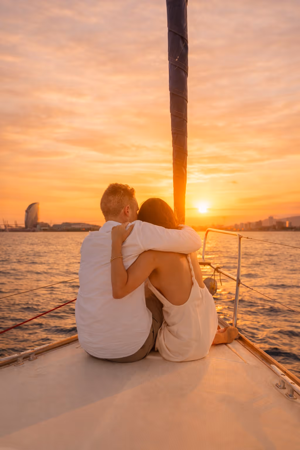 Couple embracing while sitting on the bow of a sailboat watching the sunset over the water.