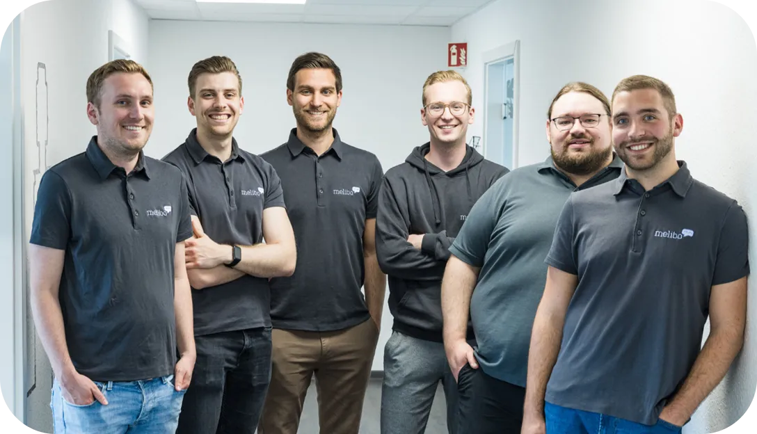 Six smiling men standing in a bright hallway wearing casual dark shirts, some with 'melibo' logos.