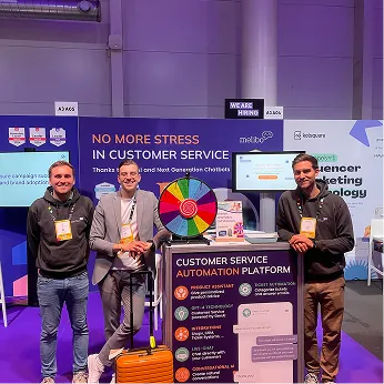 Three men standing and smiling in front of a booth with a colorful prize wheel and banner about customer service automation platform.