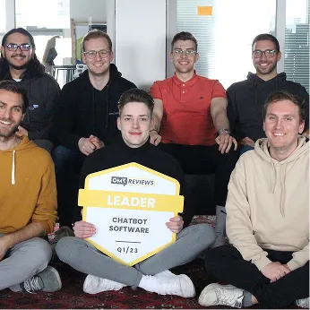 Group of six people sitting indoors smiling, with one person holding a OMR Reviews Leader Chatbot Software Q1/23 award sign.