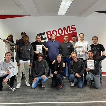 Group of 13 smiling people posing indoors in front of a wall with the word 'ROOMS', some holding certificates.
