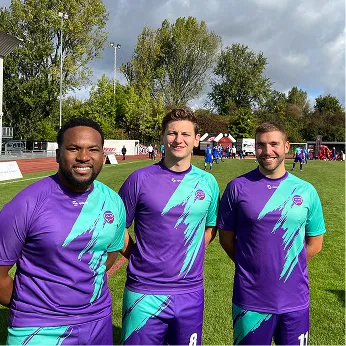 Three soccer players in purple and turquoise uniforms standing on a grass field with trees and a cloudy sky in the background.