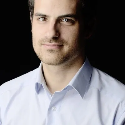 Headshot of a man in a light blue shirt with short dark hair and light facial stubble against a black background.