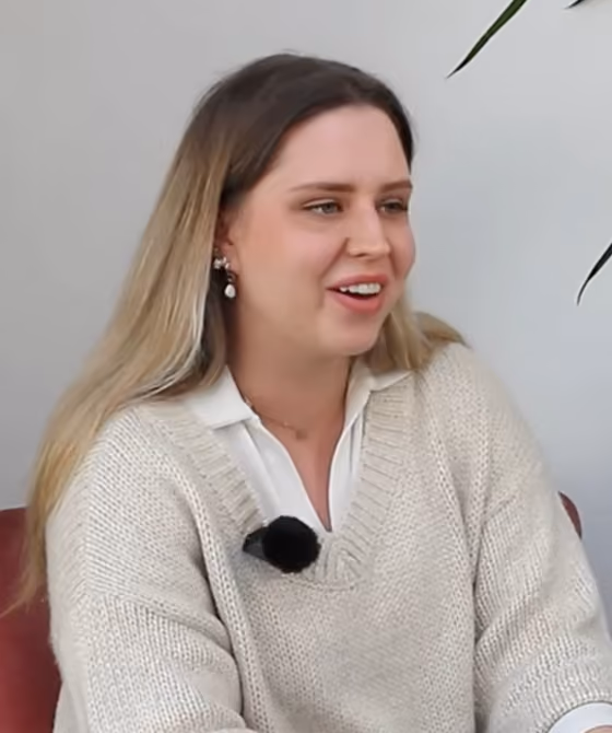 Young woman with long blonde hair wearing a cream sweater and white shirt, smiling and speaking.