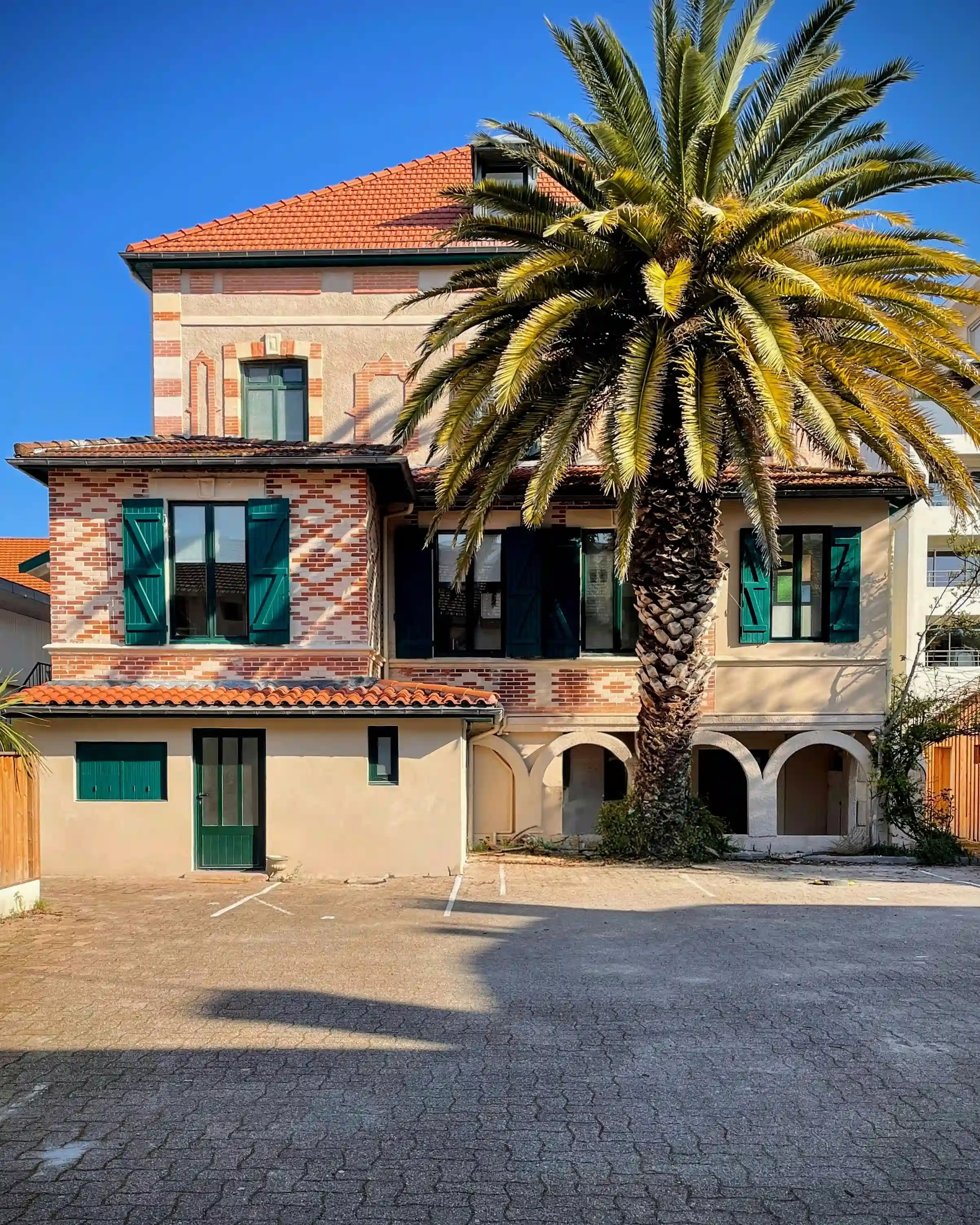 Façade de villa arcachonnaise ancienne rénovée en briques rouges, volets verts et grand palmier.