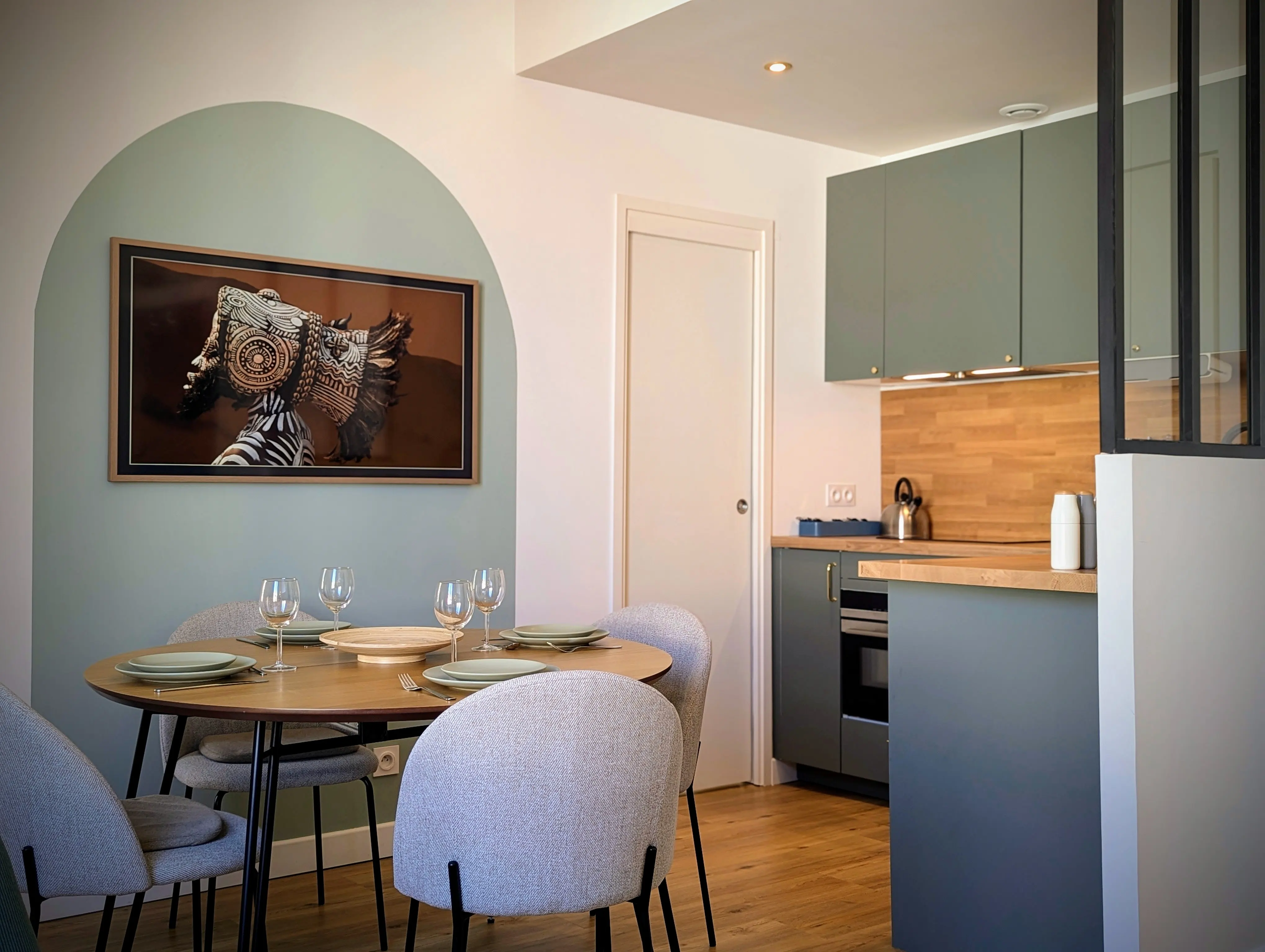 Coin repas design scandinave avec table en bois ronde, chaises grises et cuisine équipée gris-vert en arrière-plan.