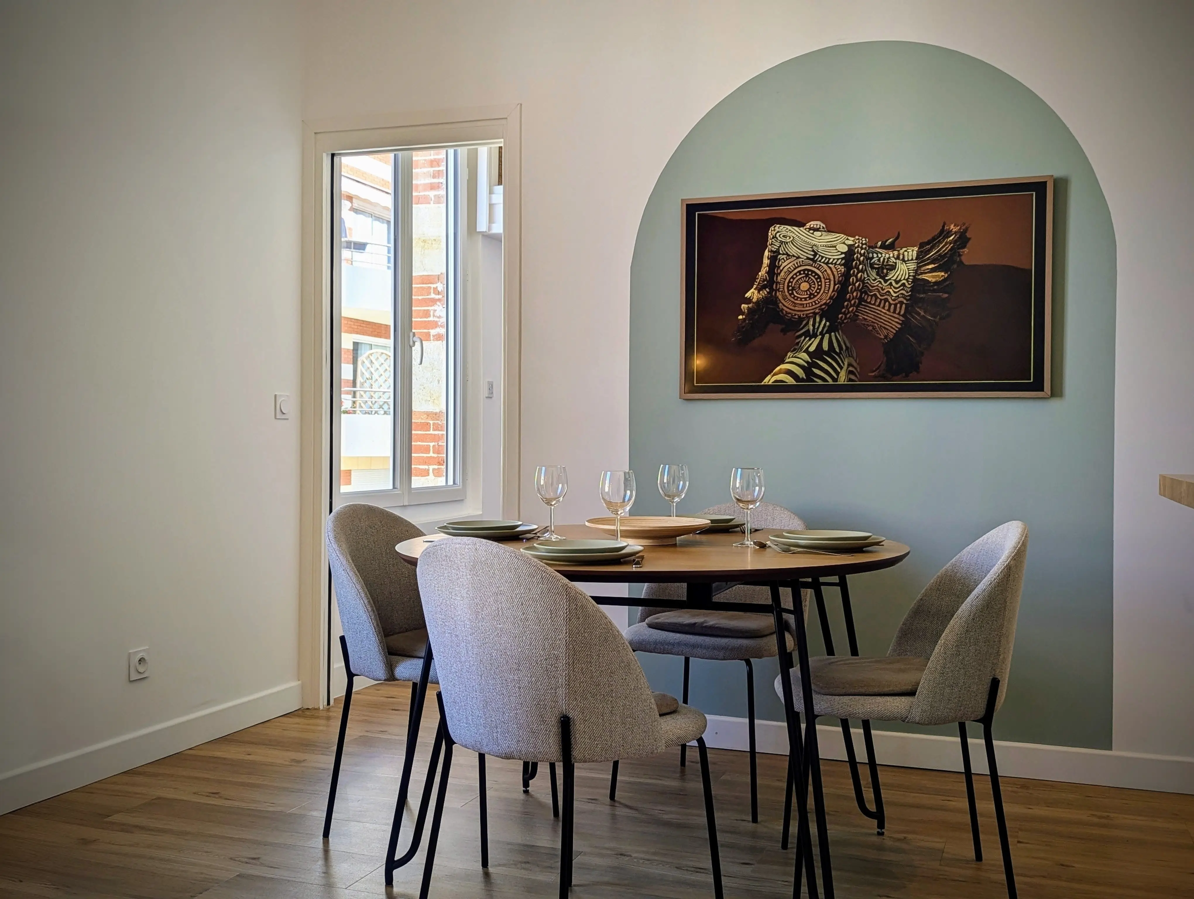 Salle à manger lumineuse avec table ronde, chaises tissu gris chiné et arche décorative verte proche d'une fenêtre.