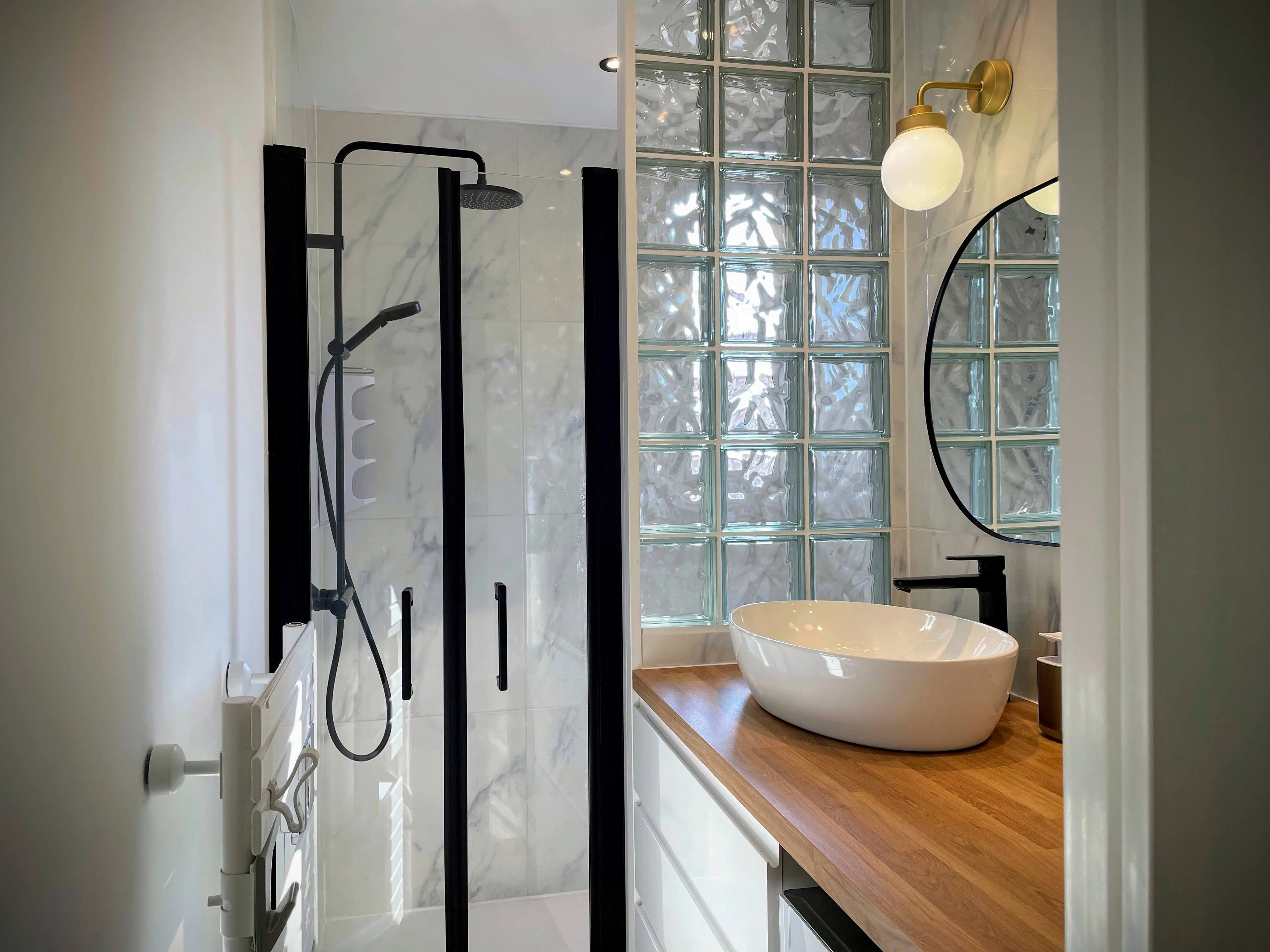 Salle de bain moderne avec paroi en pavés de verre, douche noire et meuble vasque bois.