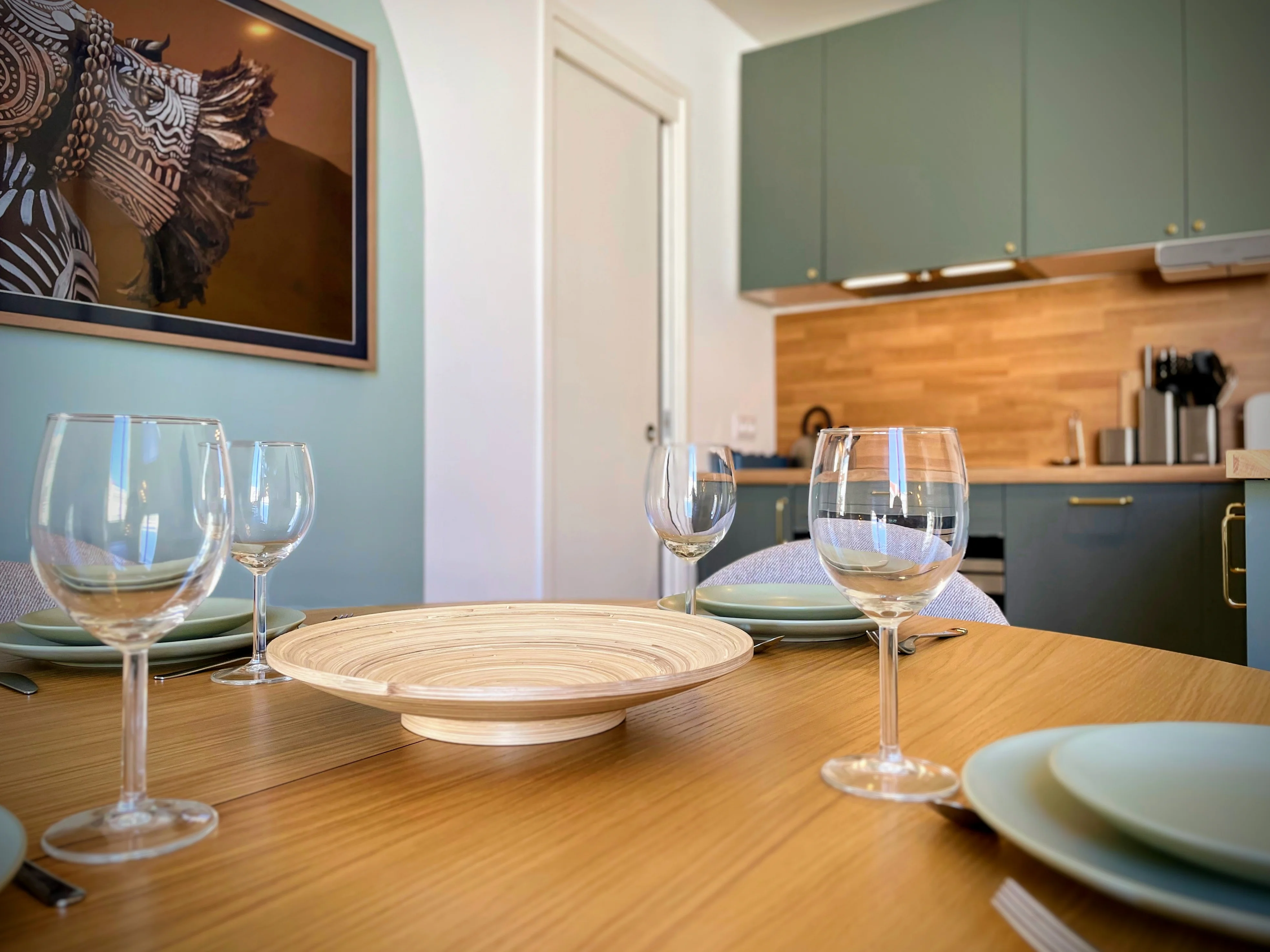 Table à manger en bois dressée avec verres à pied devant cuisine verte et bois.