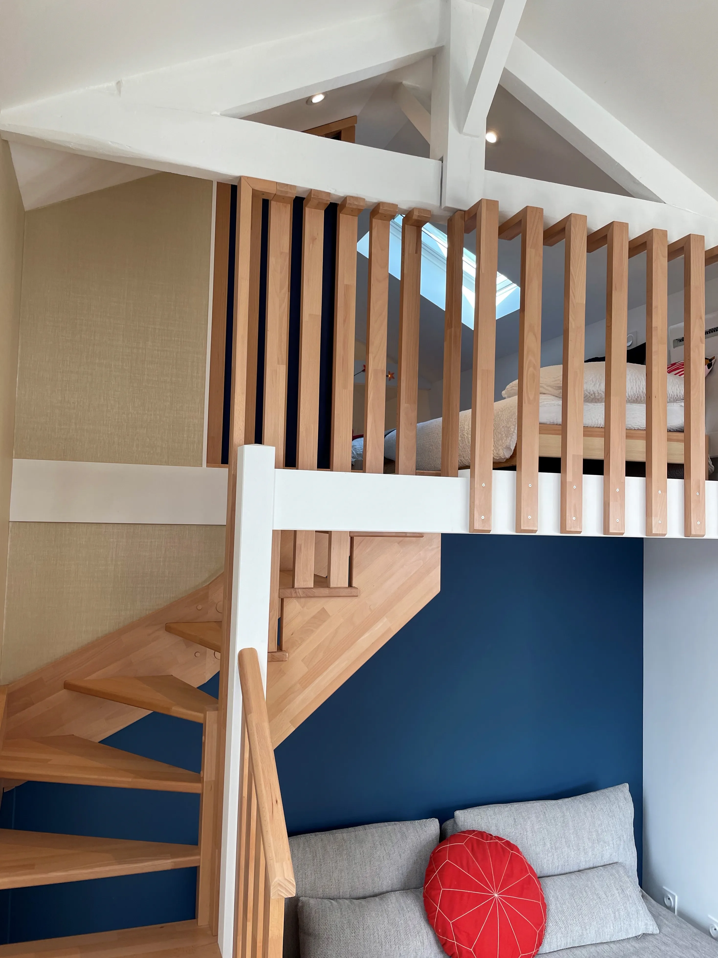 Escalier en bois menant à la mezzanine, charpente apparente blanche et garde-corps à barreaux verticaux.