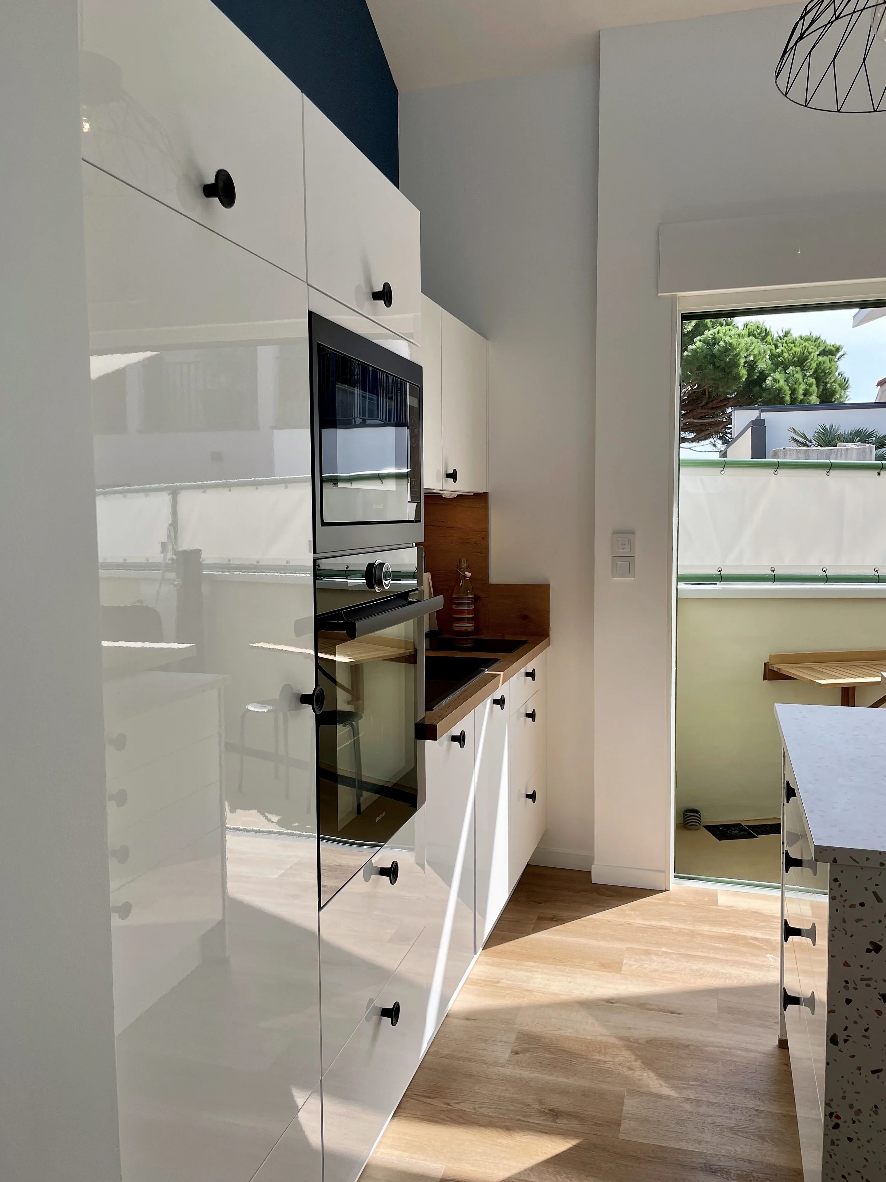 Cuisine blanche laquée avec four et micro-ondes intégrés noirs, crédence en bois et vue sur la terrasse.