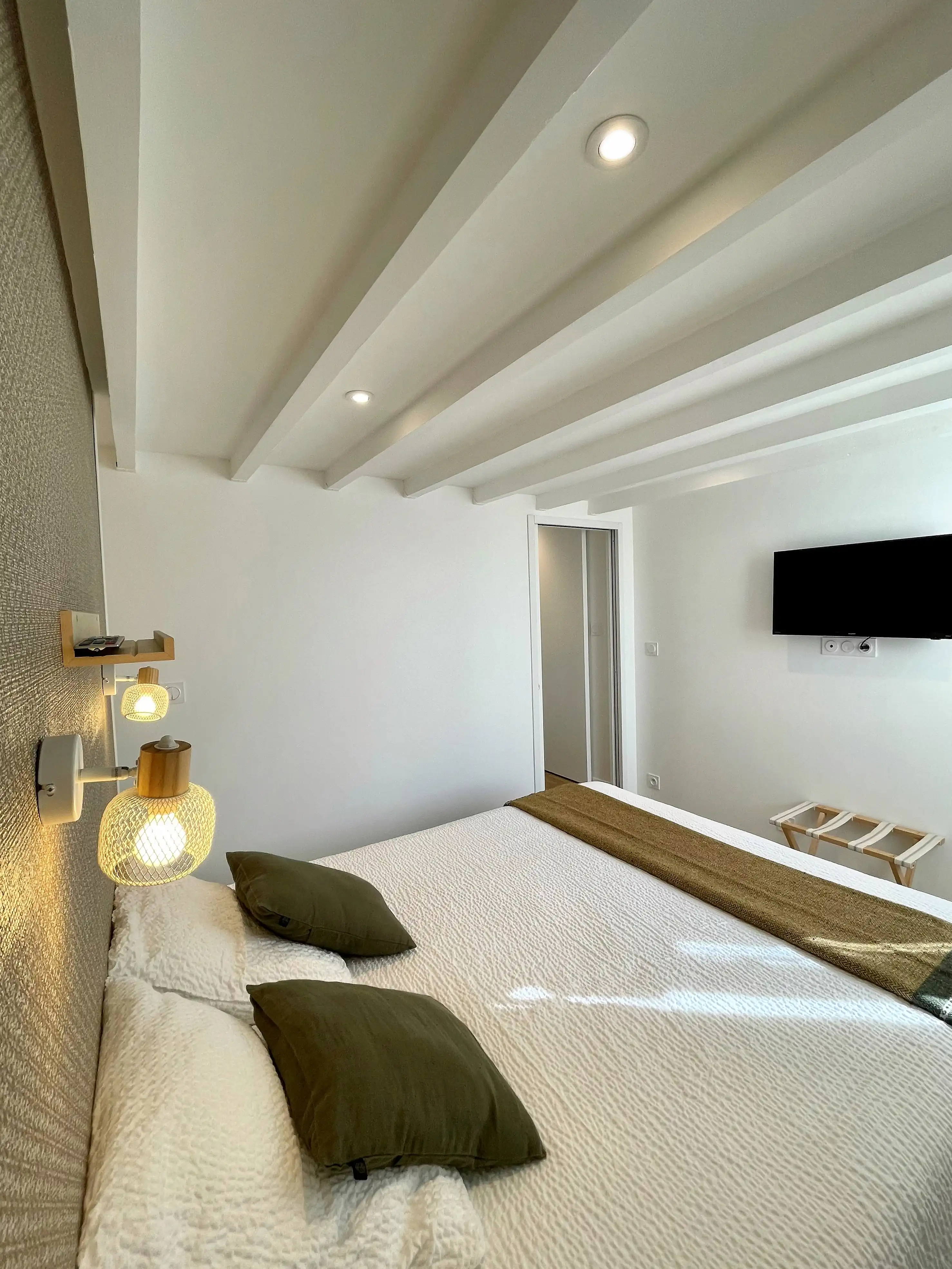Chambre sous les combles avec plafond à poutres apparentes, lit à plaid kaki et appliques murales tressées.