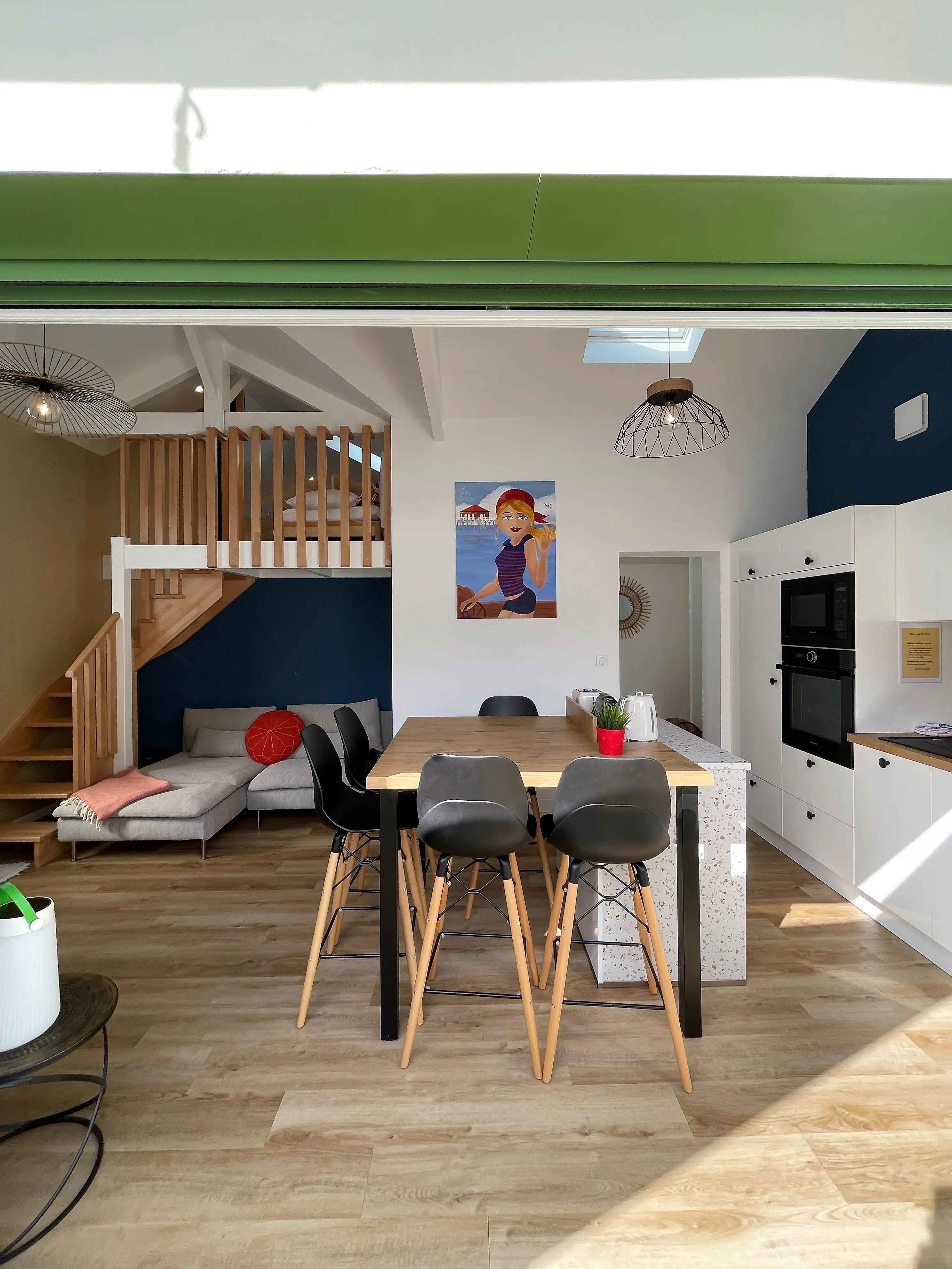 Pièce à vivre avec mezzanine en bois, mur bleu marine, canapé gris à coussin rond rouge et cuisine blanche ouverte.