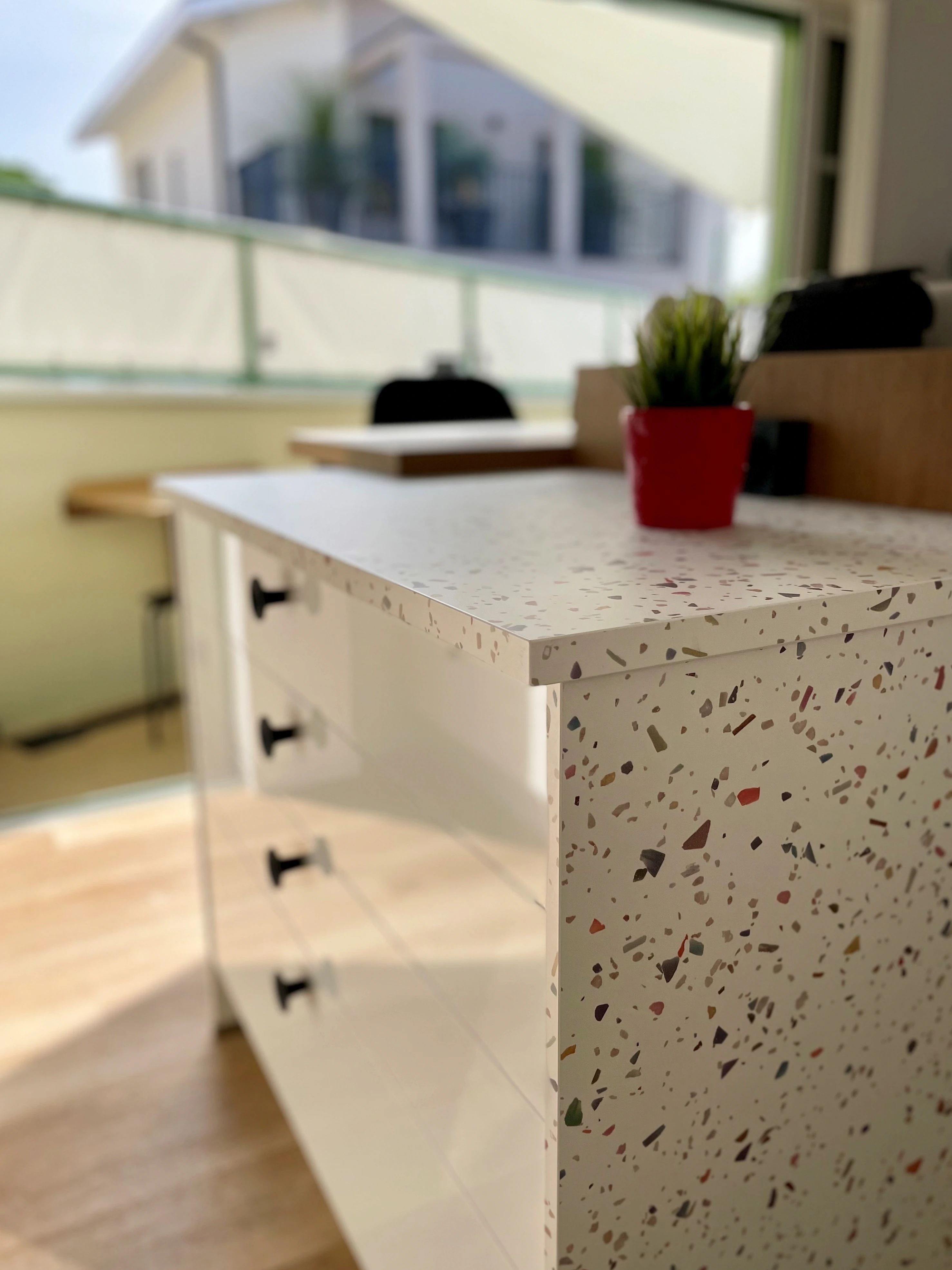 Gros plan sur un îlot de cuisine en terrazzo blanc à tiroirs noirs, pot de fleur rouge et terrasse en fond.