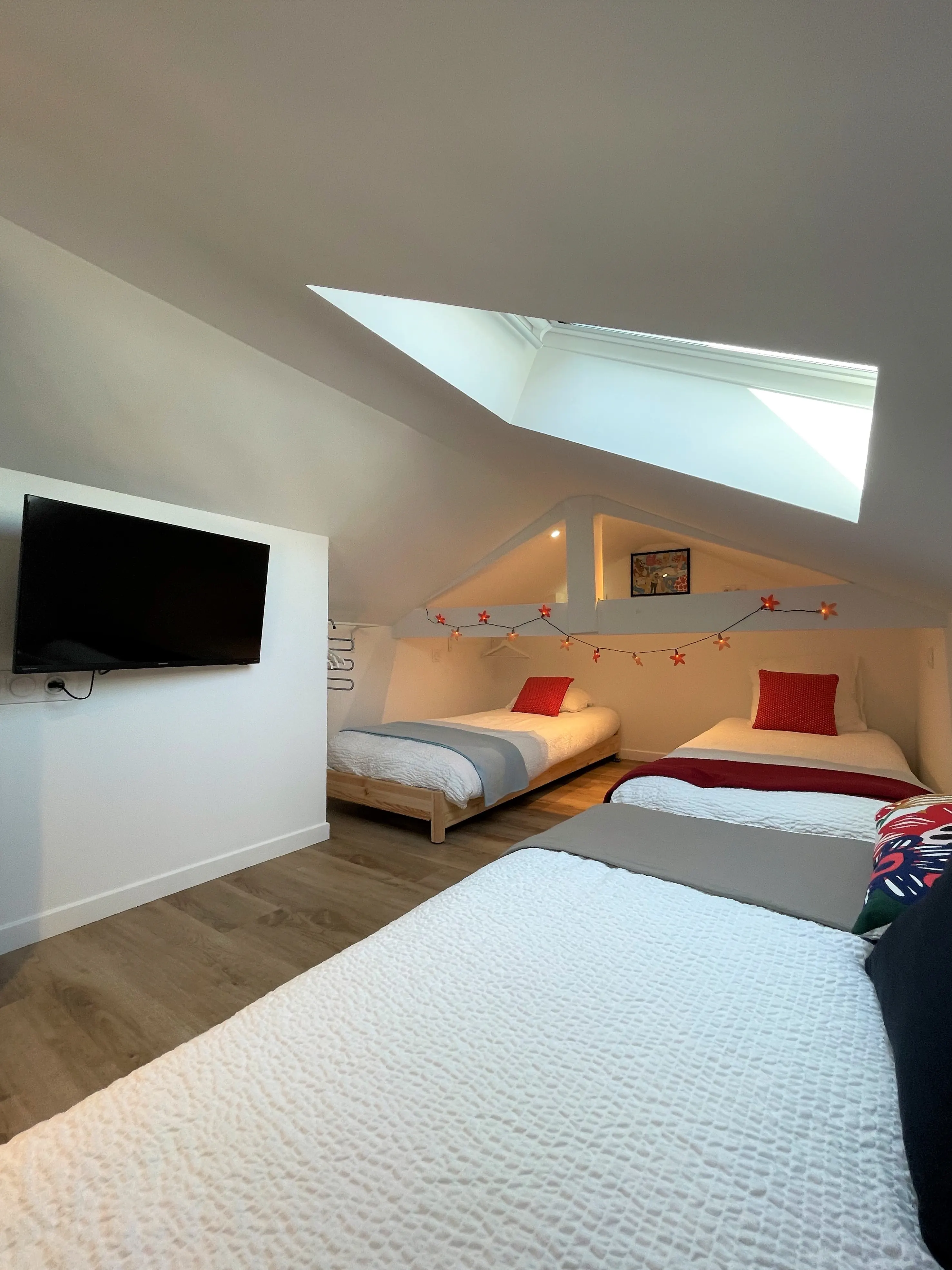 Chambre sous les combles avec trois lits, fenêtre de toit et guirlande lumineuse en forme de fleurs.