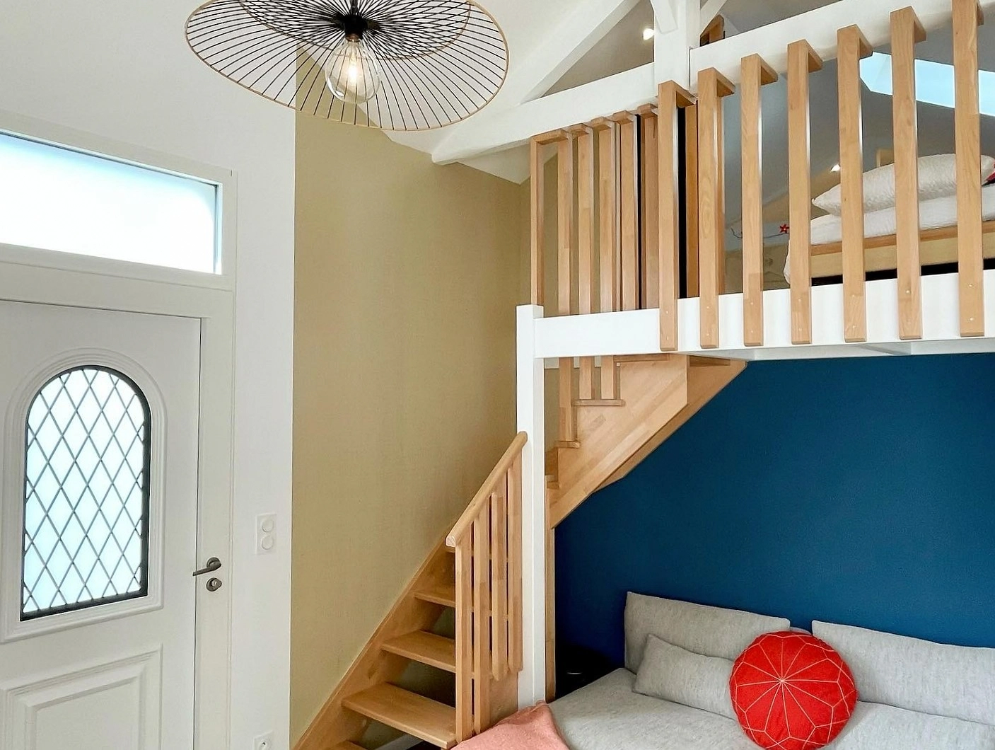Entrée avec porte vitrée à losanges, suspension tressée, escalier vers la mezzanine et canapé gris.