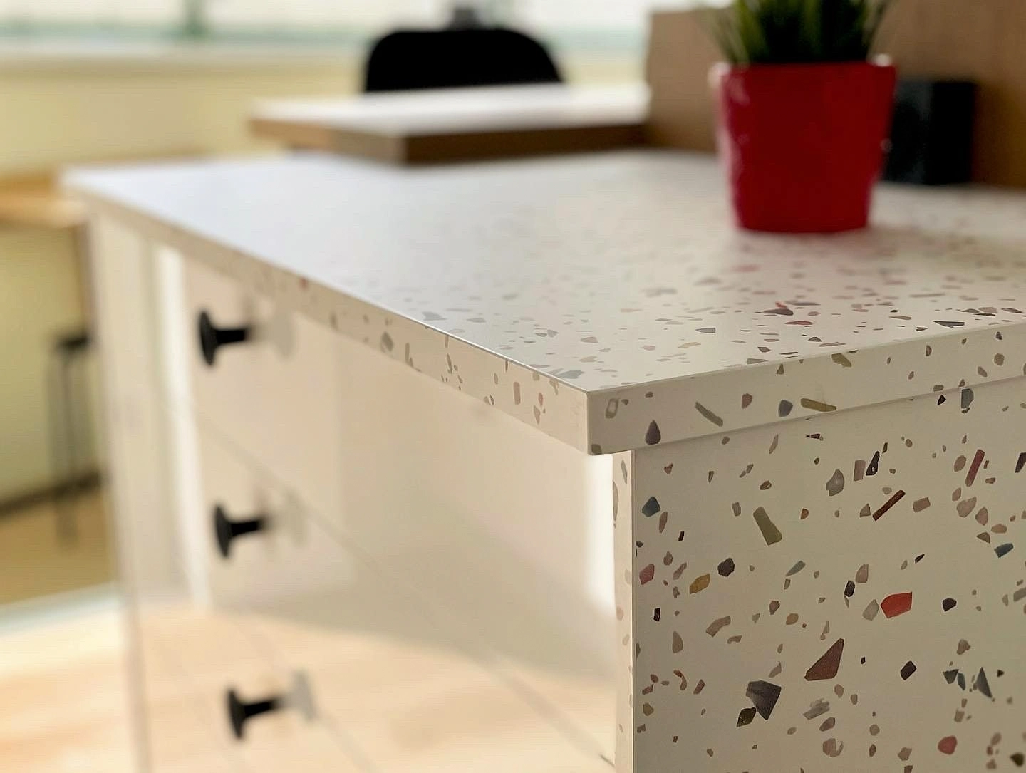 Gros plan sur l'angle d'un îlot de cuisine en terrazzo blanc, pot de fleur rouge en arrière-plan.