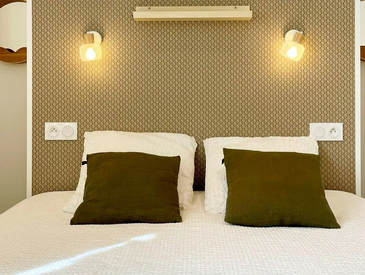 Tête de lit avec coussins kaki sur tissu texturé doré, appliques tressées allumées de chaque côté.