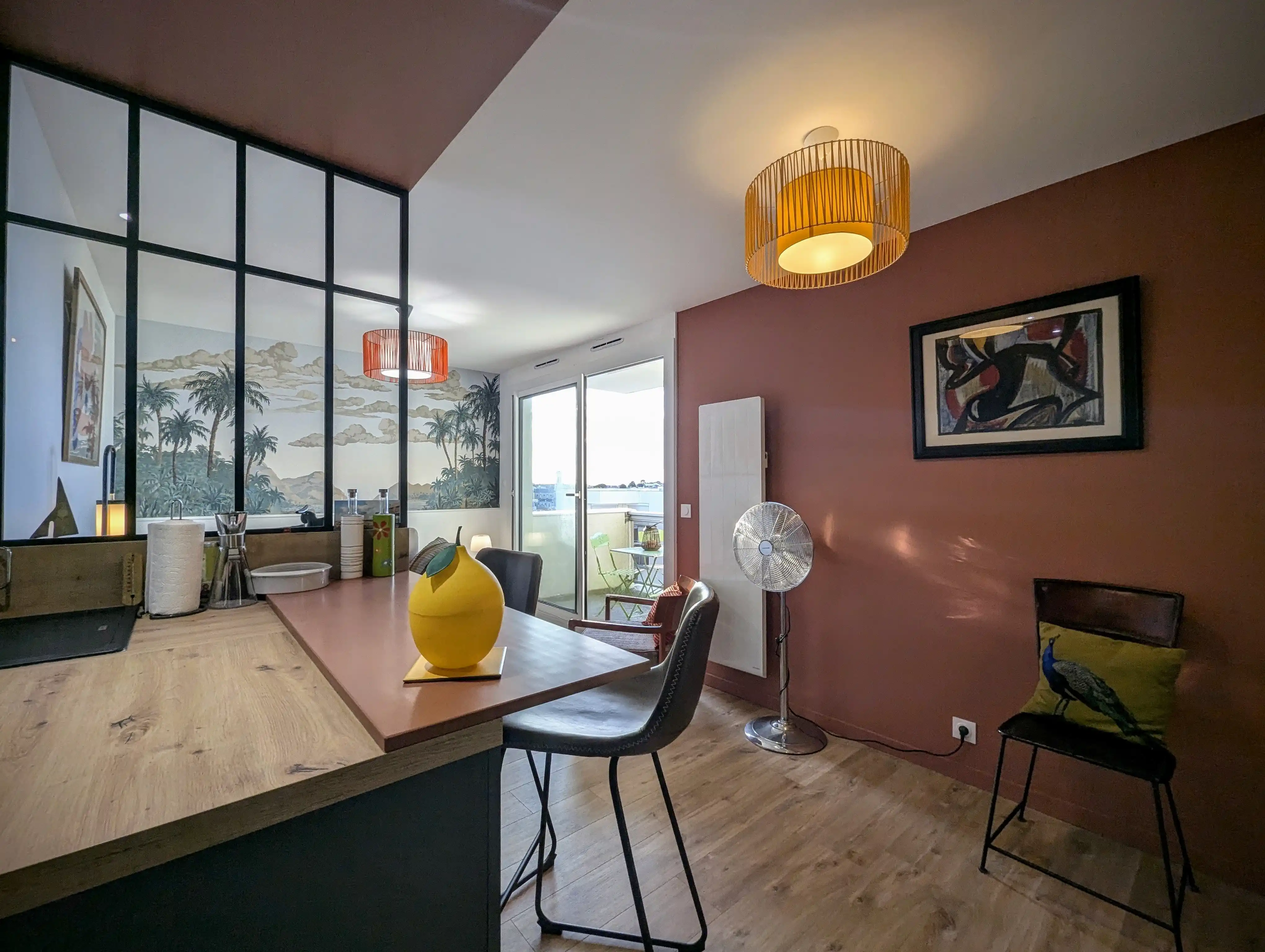 vue de la partie séjour depuis la cuisine d'un studio rénové sur Arcachon, papier peint panoramique Palmier Impérial, baie atelier pour qui sépare l'espace cuisine avec meubles bleu velouté. Murs terracotta.