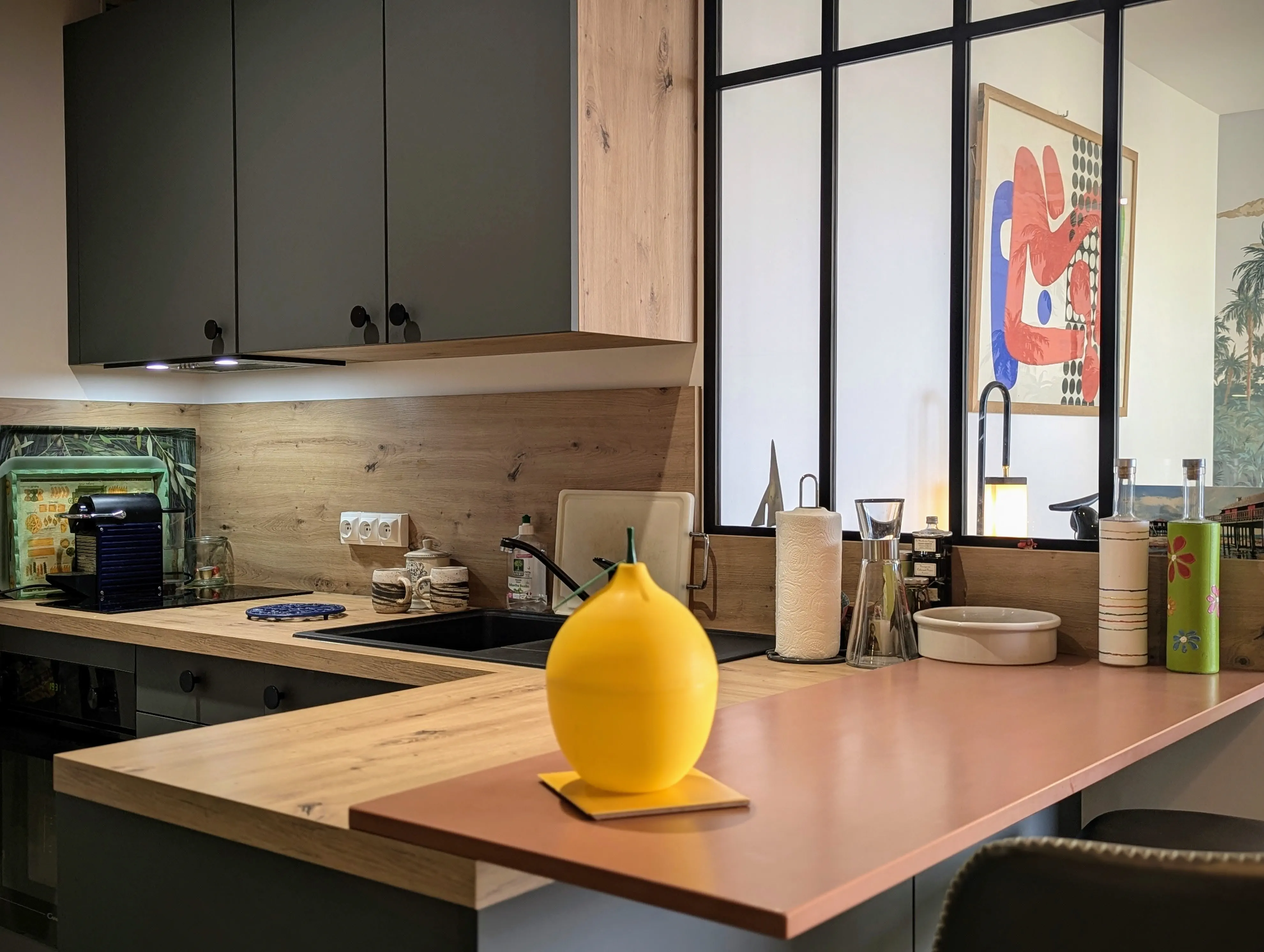 vue de la partie cuisine d'un studio rénové sur Arcachon, papier peint panoramique Palmier Impérial, baie atelier pour qui sépare l'espace cuisine avec meubles bleu velouté. Plan de travail effet bois Murs terracotta.