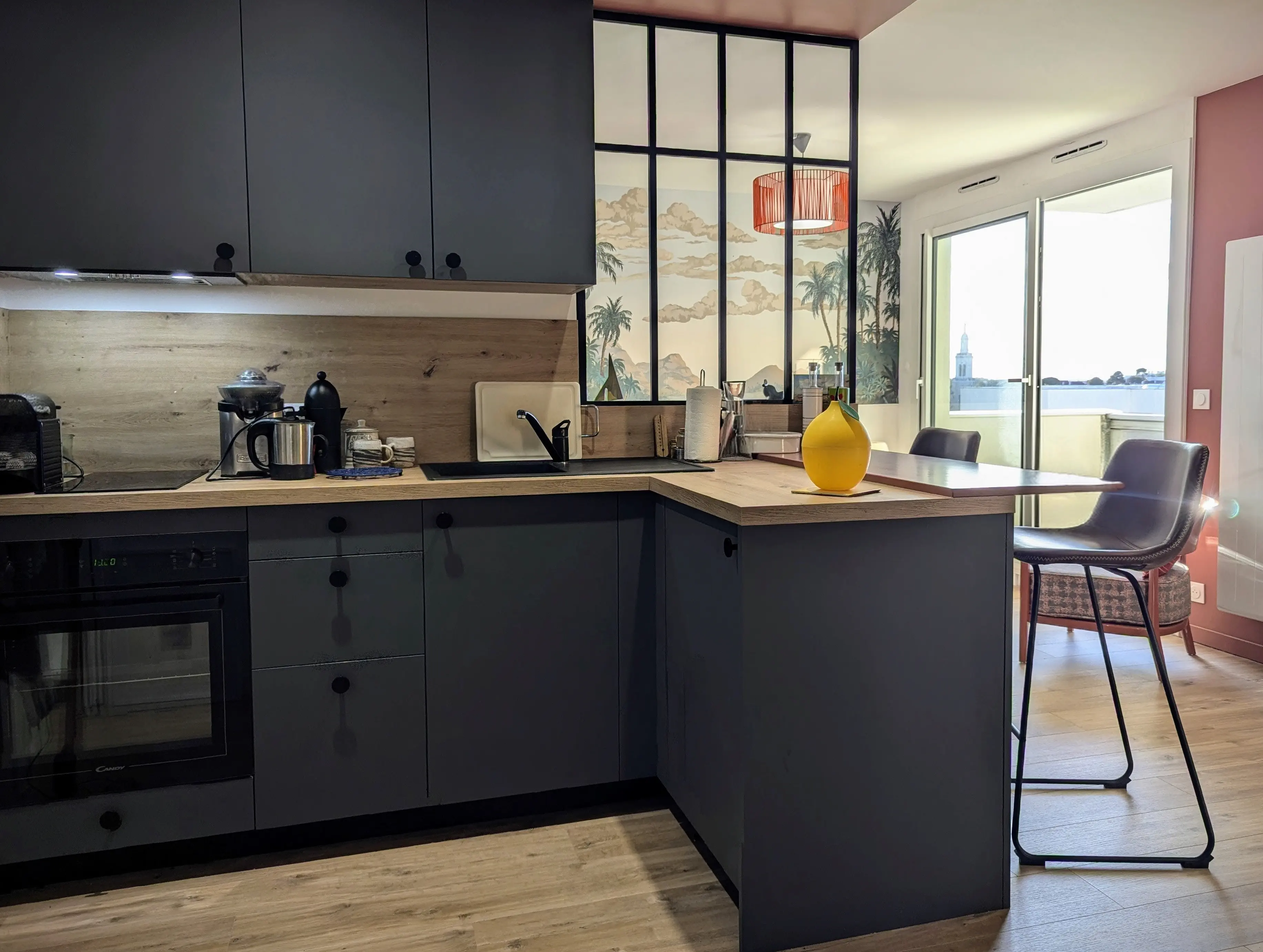 vue de la partie cuisine d'un studio rénové sur Arcachon, papier peint panoramique Palmier Impérial, baie atelier pour qui sépare l'espace cuisine avec meubles bleu velouté. Plan de travail effet bois Murs terracotta.