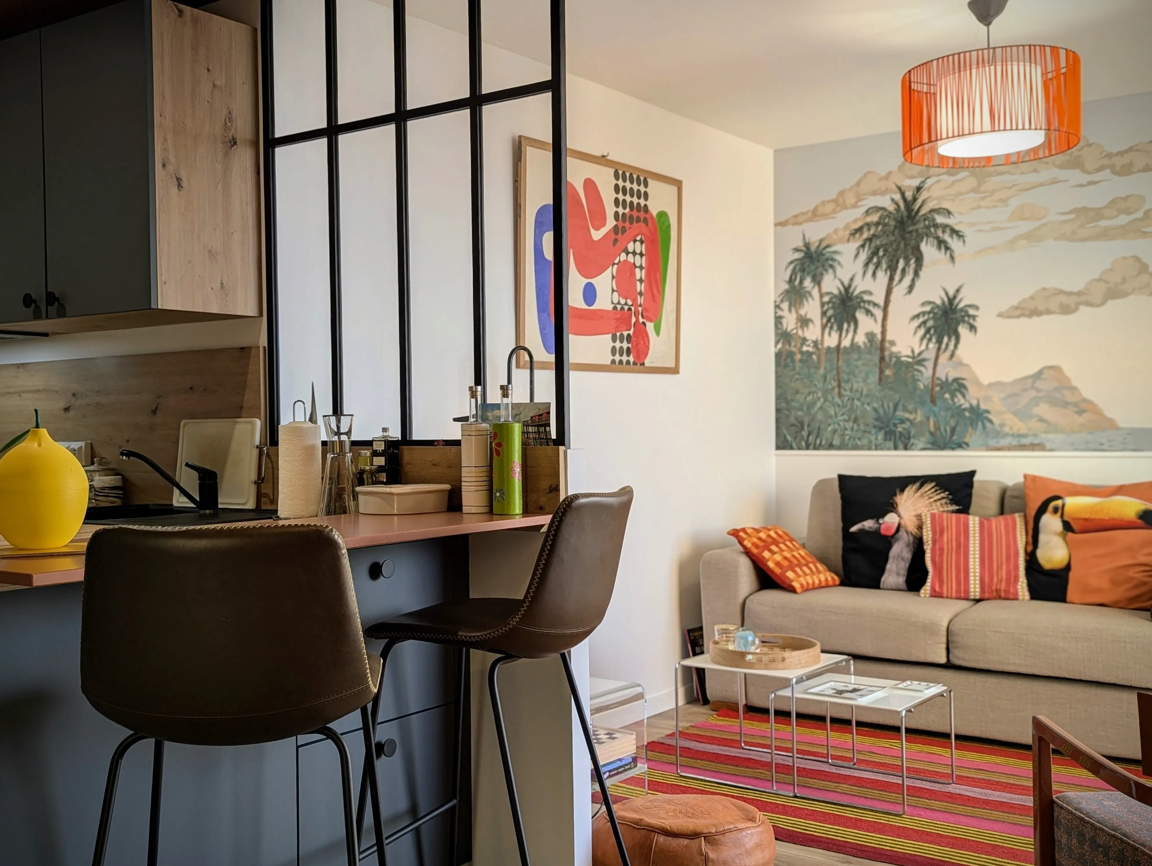 vue de la partie séjour depuis la cuisine d'un studio rénové sur Arcachon, papier peint panoramique Palmier Impérial, baie atelier pour qui sépare l'espace cuisine avec meubles bleu velouté.
