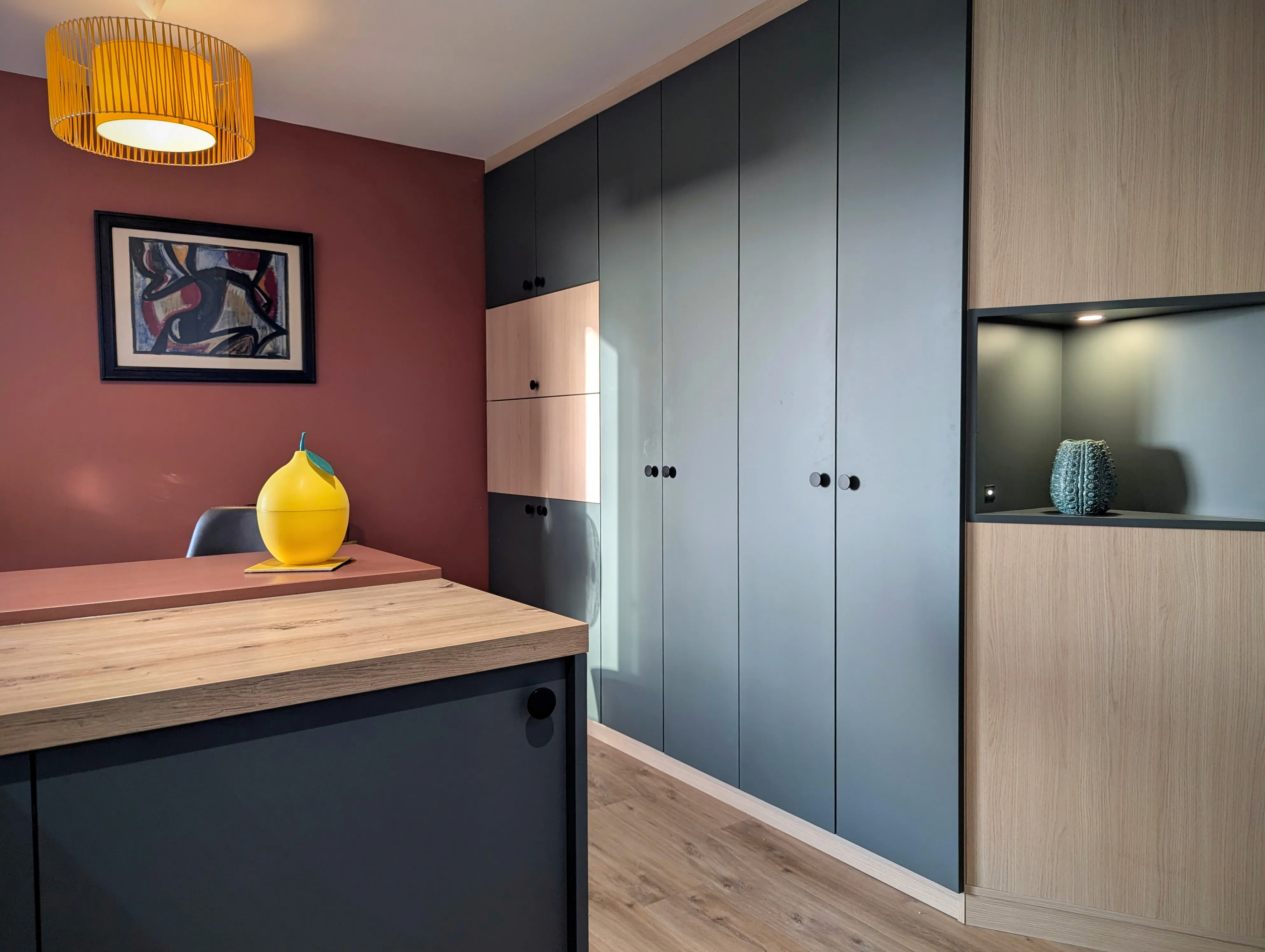 studio rénové sur Arcachon, meuble sur mesure dans des tons bois et vert avec secrétaire intégré, baie atelier pour qui sépare l'espace cuisine avec meubles bleu velouté. Murs terracotta.
