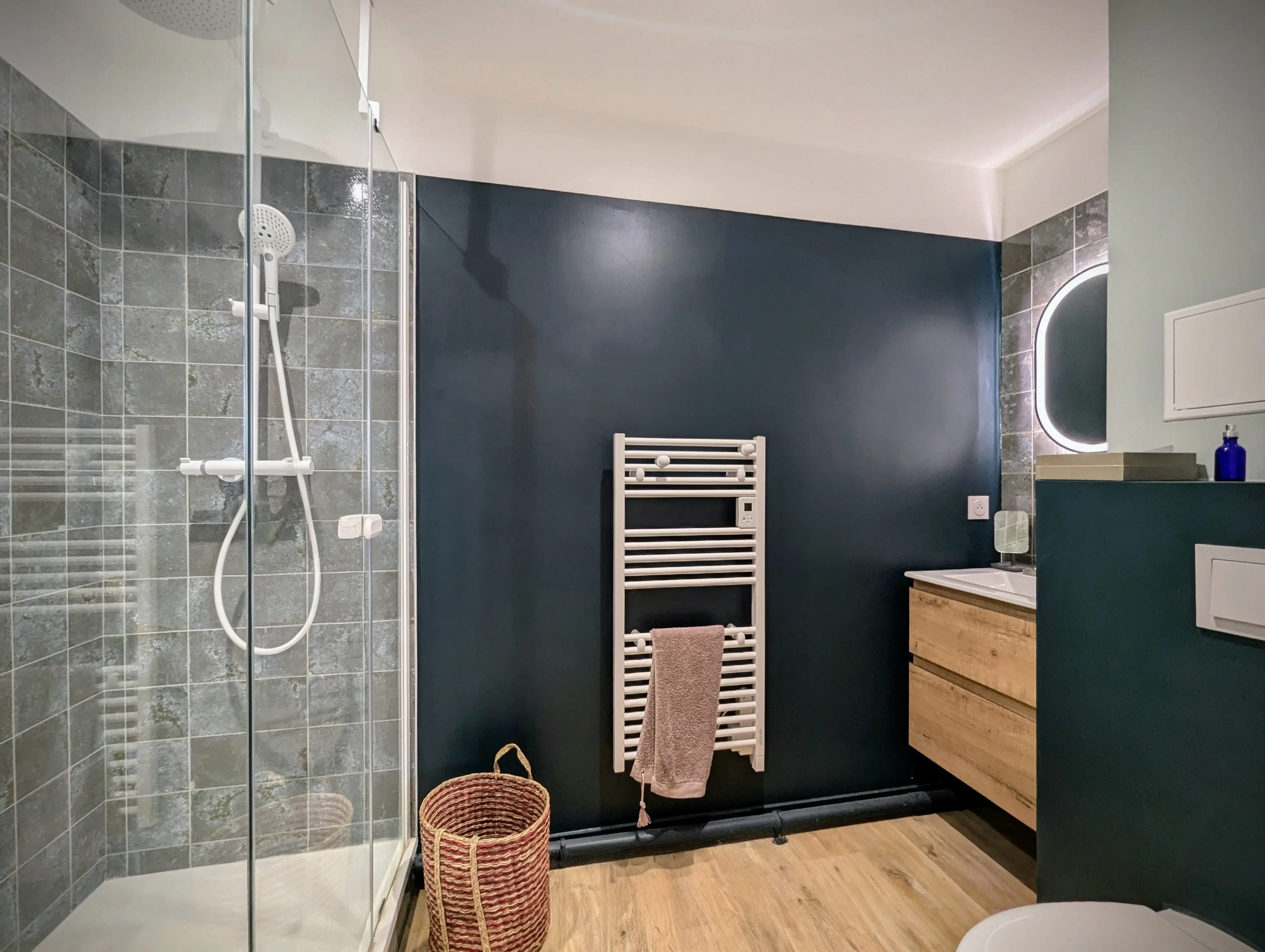 vue d'une salle d'eau moderne avec meuble vasque. Miroir rétroéclairé. Carrelage mural en terre cuite. Murs bicolore vert d'eau et vert foncé. Mur en bleu velouté. Sol vinyle effet chêne. Douche bac extra plat blanc.