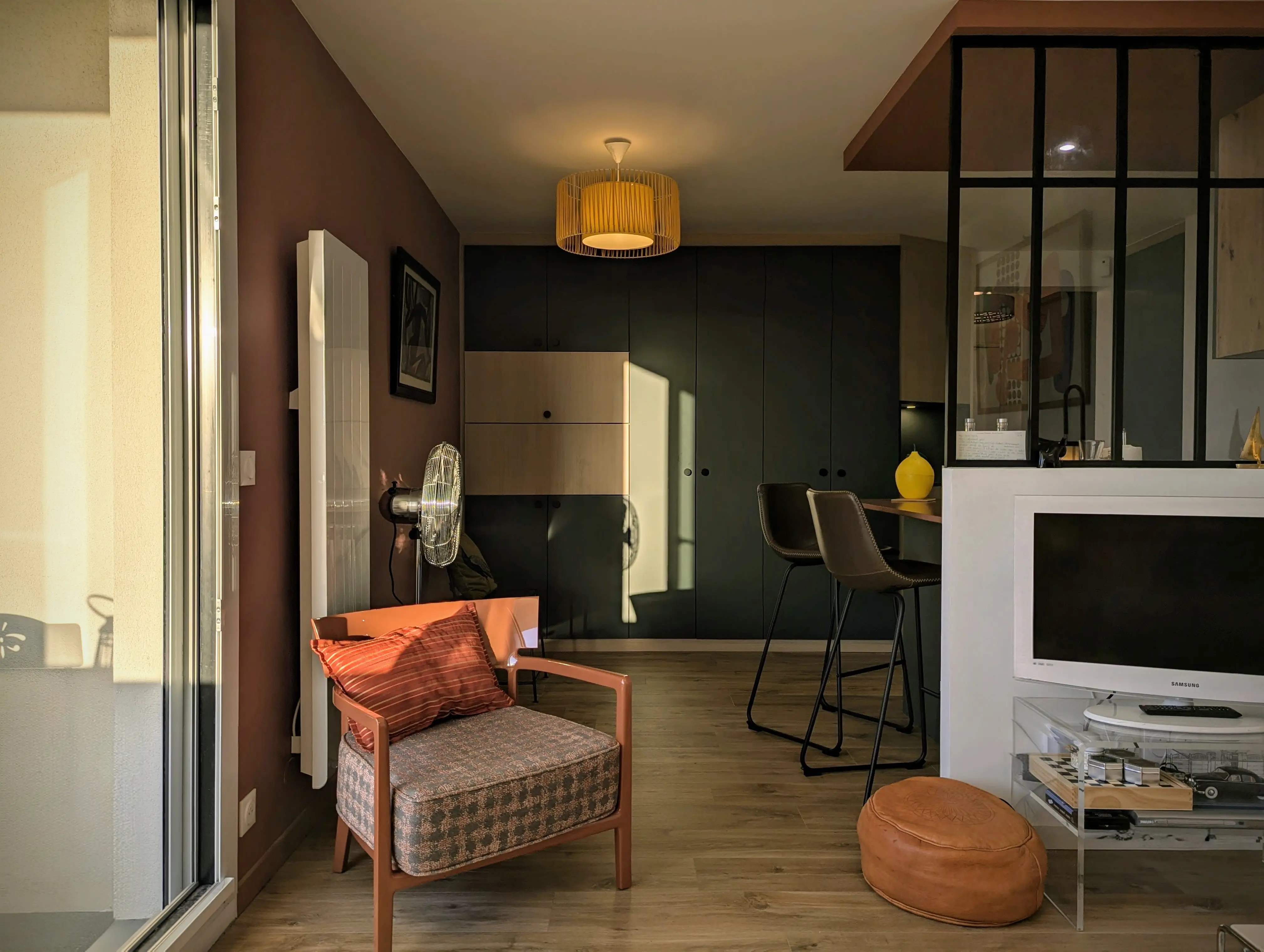 studio rénové sur Arcachon, meuble sur mesure dans des tons bois et vert avec secrétaire intégré, baie atelier pour qui sépare l'espace cuisine avec meubles bleu velouté. Murs terracotta.