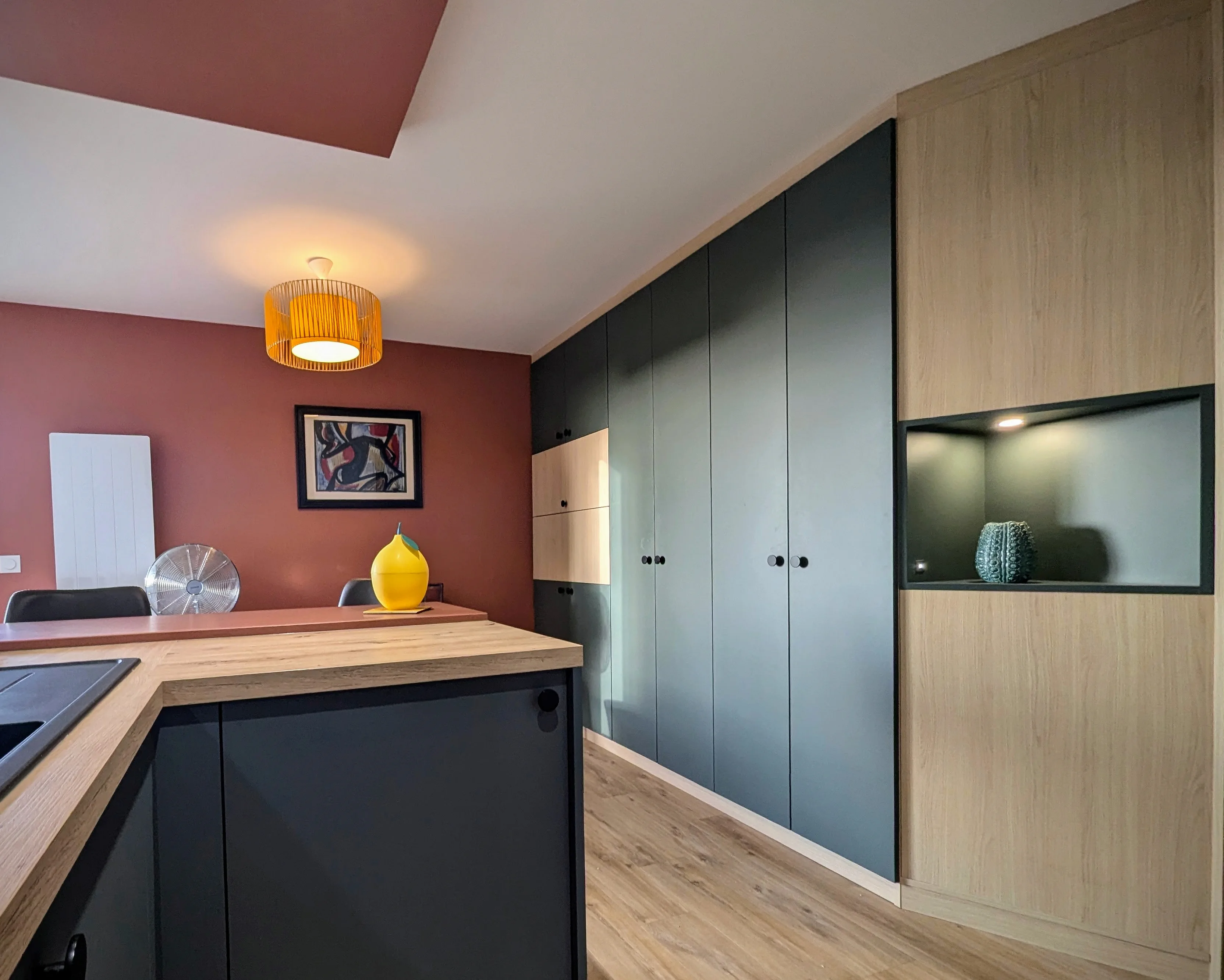 studio rénové sur Arcachon, meuble sur mesure dans des tons bois et vert avec secrétaire intégré, baie atelier pour qui sépare l'espace cuisine avec meubles bleu velouté. Murs terracotta.