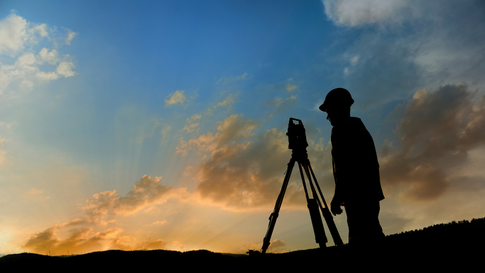A silhouette of a person standing in front of a surveyor equipment at sunset